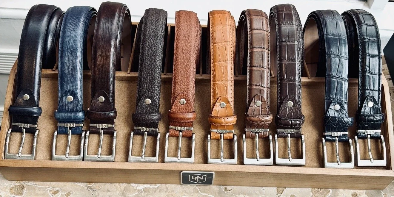 An assortment of Scarborough custom men's leather alligator belts in various colors arranged on a wooden display stand.