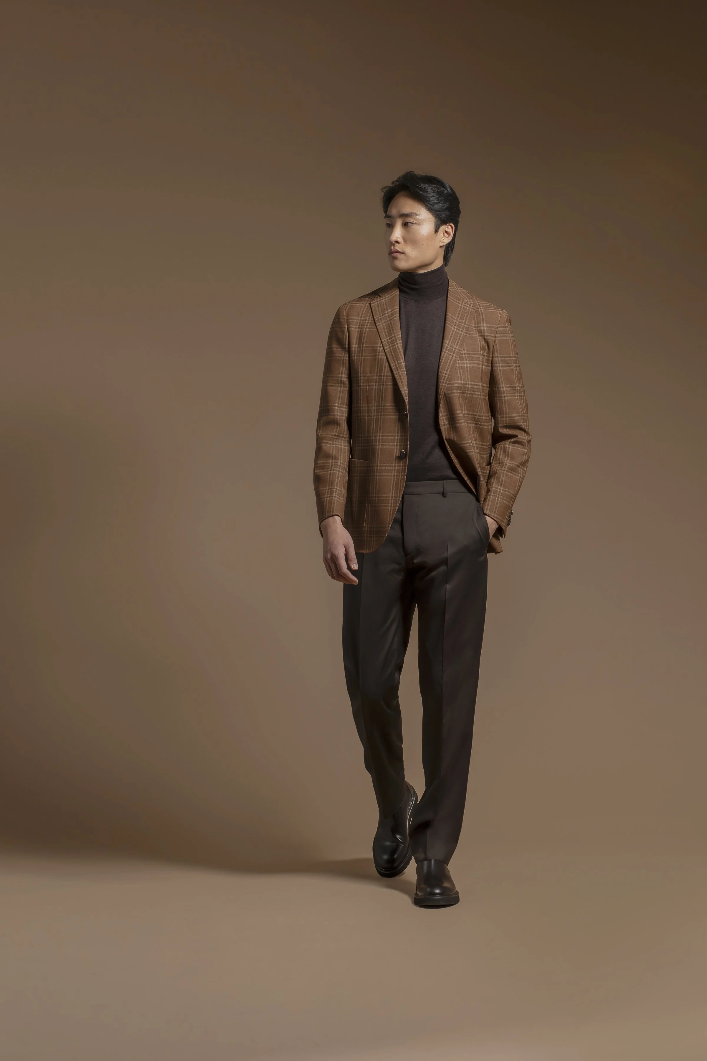 A man in a Scabal brown plaid blazer, dark turtleneck, gray dress pants, and black shoes walking, against a plain brown background.