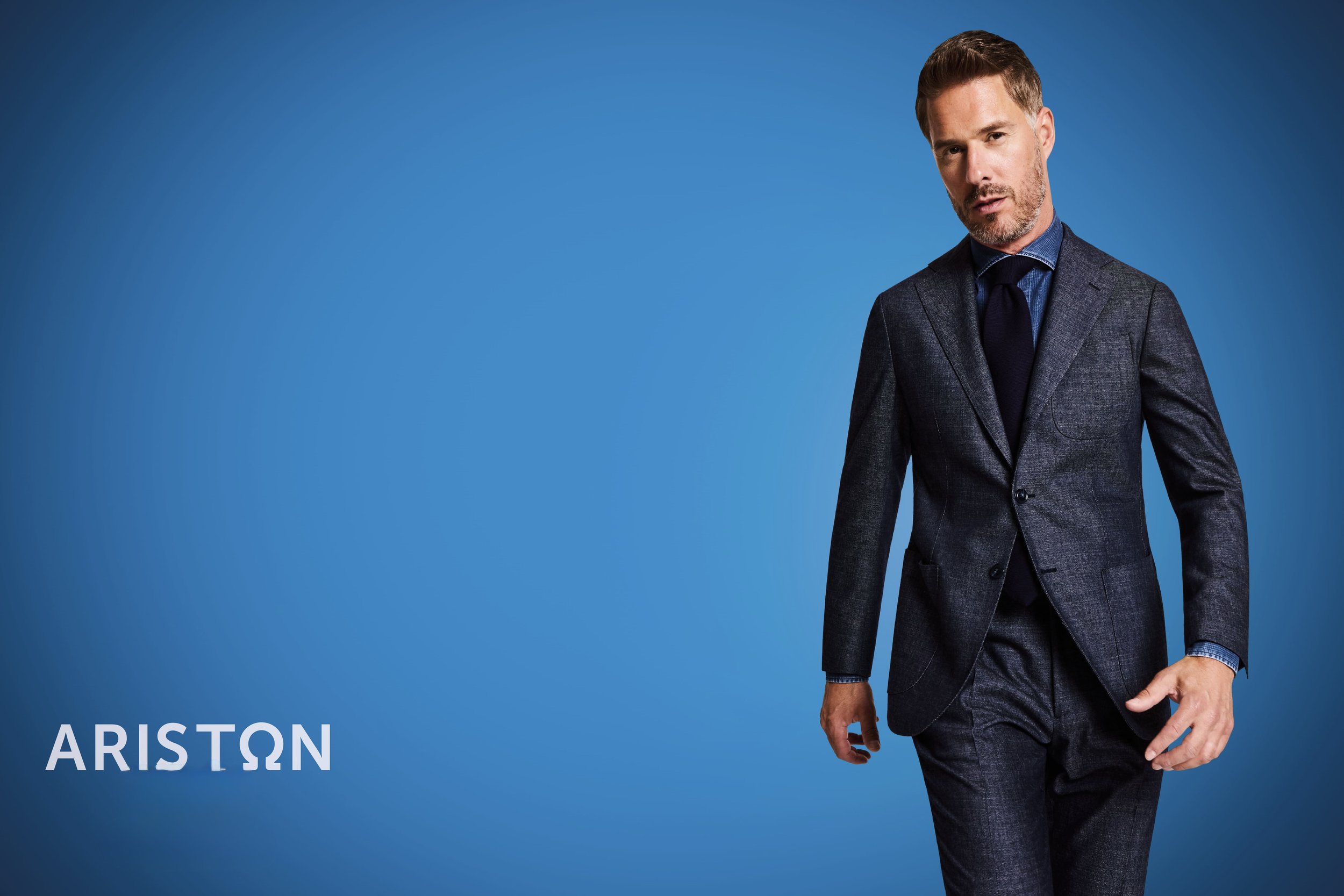 A man wearing a dark suit with a blue shirt and black tie posing against a blue background, with the brand name Ariston displayed in the lower left corner.