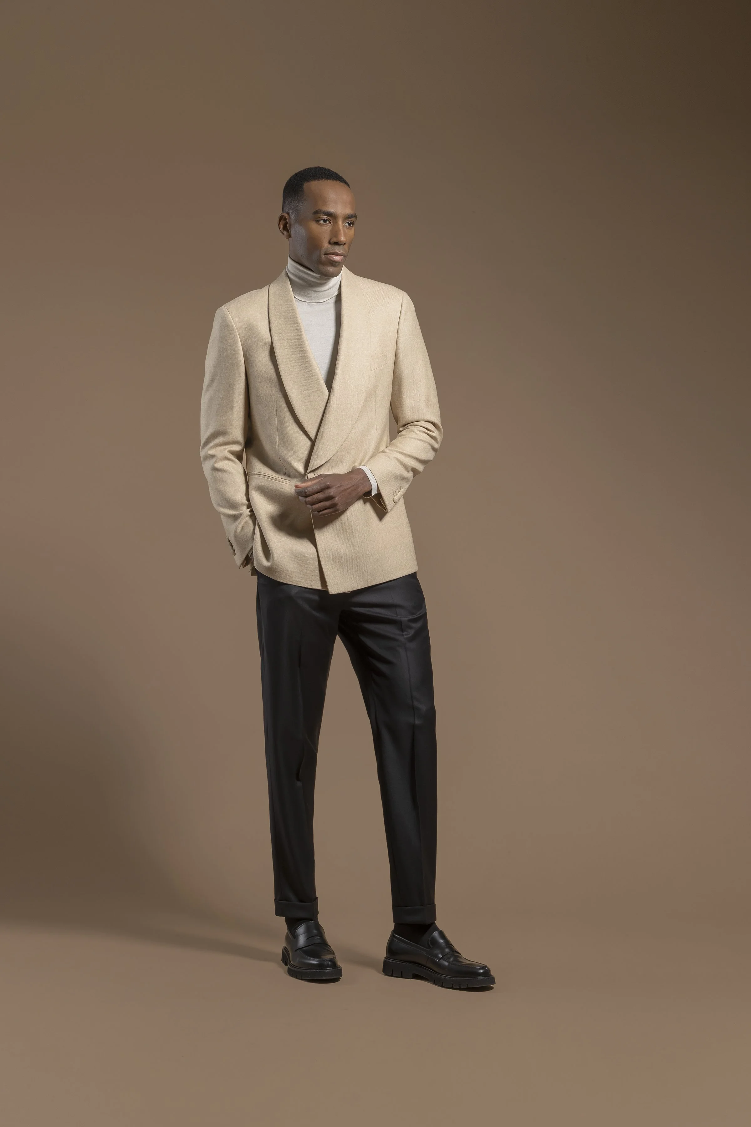 A man wearing a Scabal beige blazer over a white turtleneck, black pants, and black boots standing against a plain brown background.