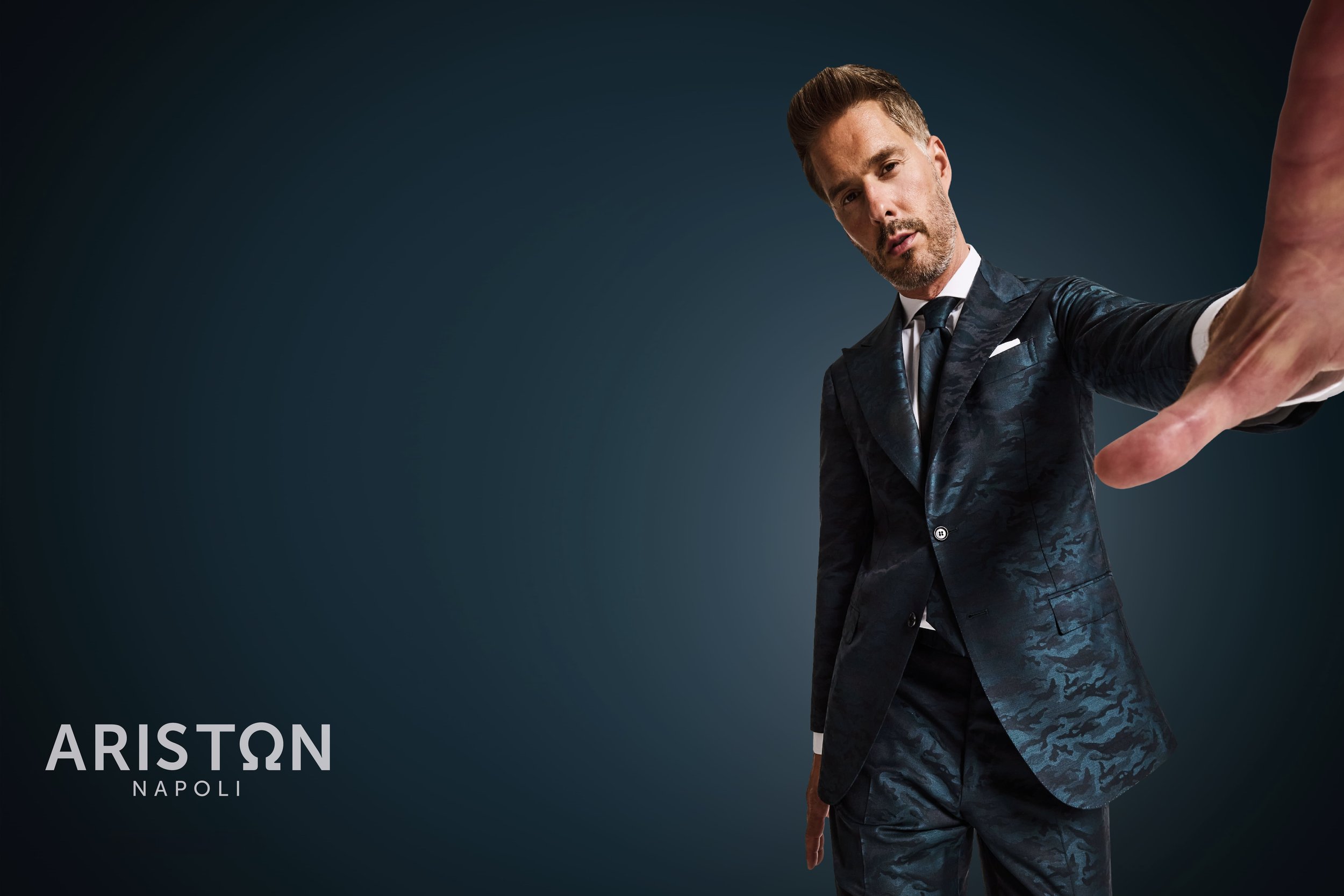 A man in a dark blue suit takes a selfie with a serious expression, extending his arm toward the camera, against a dark background. The text 'ARISTON NAPOLI' appears in the lower left corner.