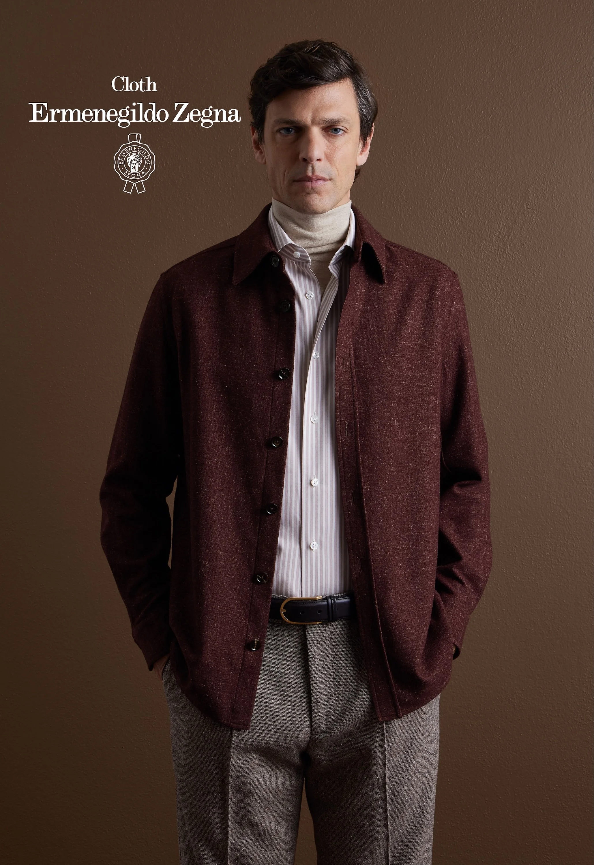 A man is standing against a brown wall, wearing a brown Zegna coat, a cream turtleneck, a white striped shirt, and gray wool pants.