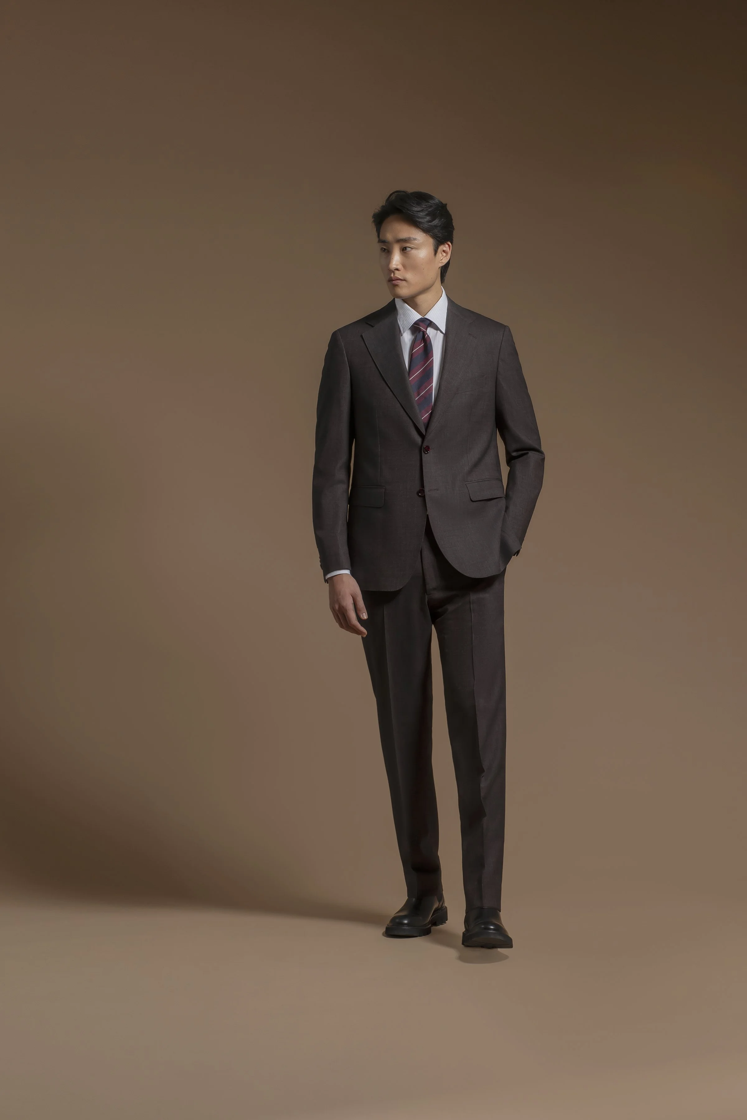 A man standing against a plain brown background, wearing a Scabal dark suit, white shirt, striped tie, and black shoes, with one hand in his pocket, looking slightly to his left.