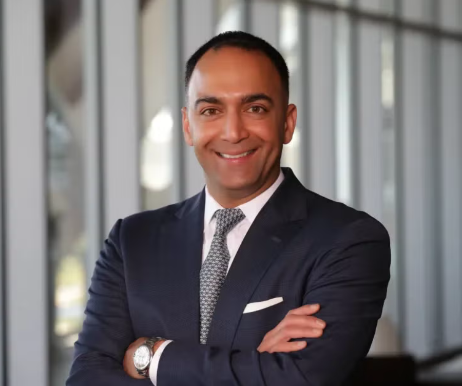 A man in a dark suit and patterned tie standing in front of a windowed background, smiling with his arms crossed.