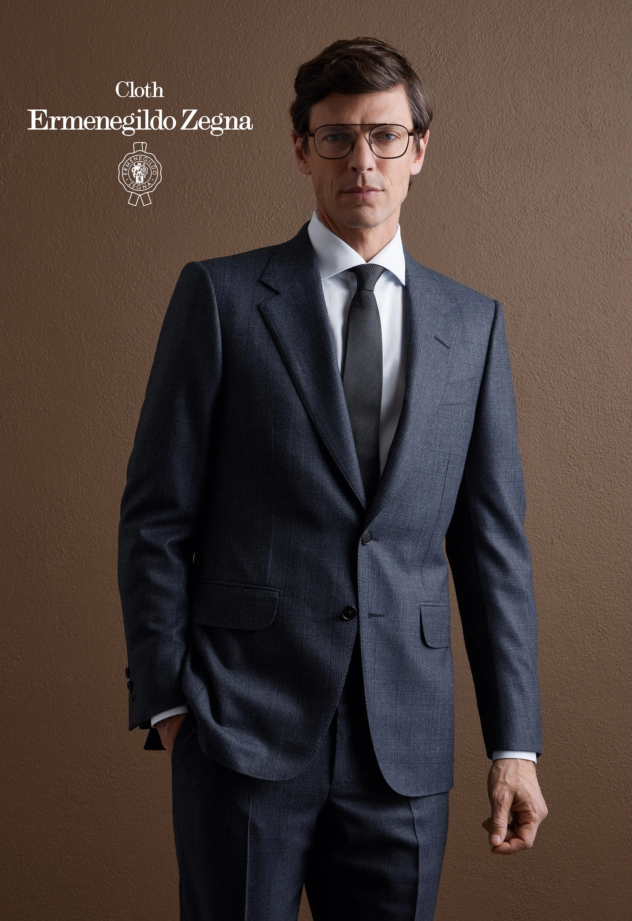 A man in a gray Zegna suit, white shirt, and black tie standing against a brown wall, wearing glasses with a serious expression.
