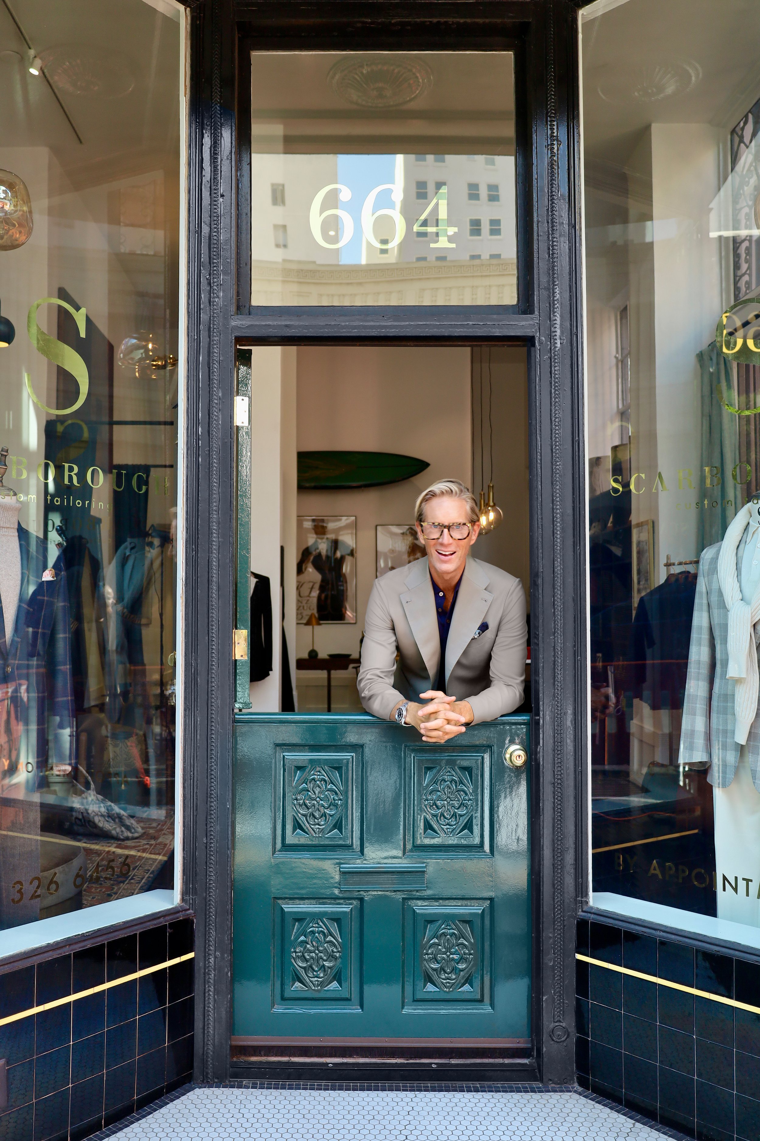 Christian with glasses and a light gray blazer leaning on a green door frame at the entrance of Scarborough Custom Tailoring, smiling. The store's address 664 is visible on the glass above the door, with mannequins and clothing displays inside.
