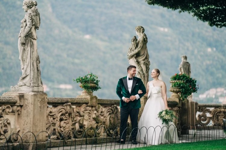 A bride and groom wearing Scarborough Custom Tailoring tuxedo stand on a stone terrace with ornate statues and potted plants, smiling at each other during their wedding photo shoot.