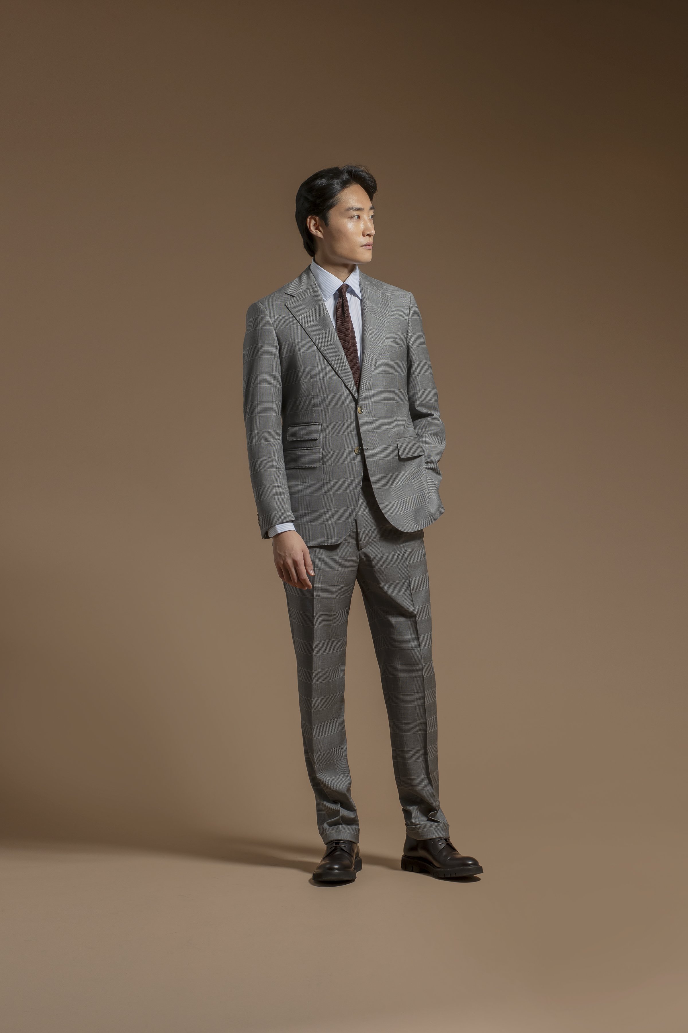 A man standing in a studio with a plain brown background, wearing a Scabal gray plaid suit, white shirt, brown tie, and black boots, looking to the side.