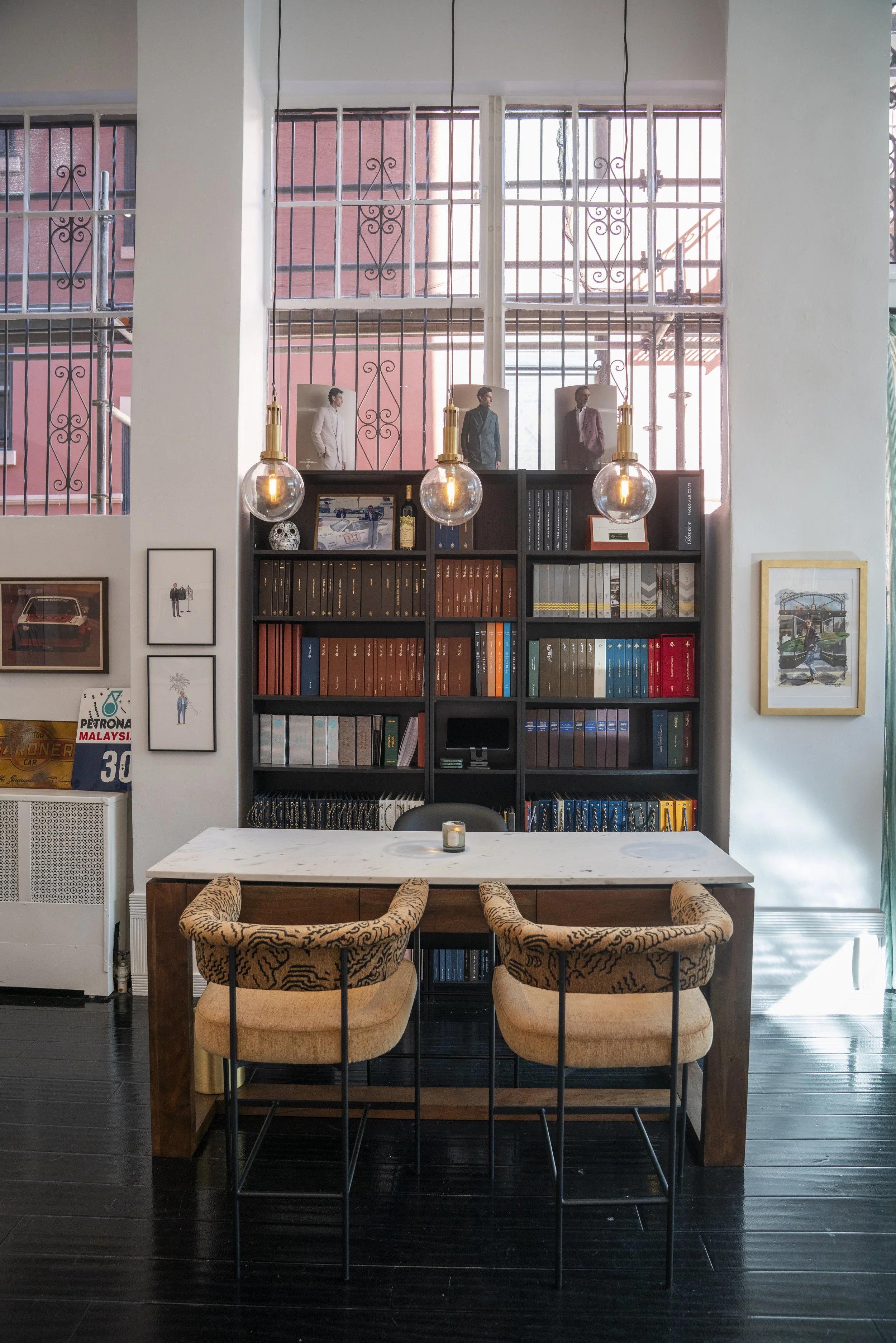 Interior of the work area with a marble-topped table, two chairs with leopard print cushions, black flooring, a black bookshelf filled with books and decor, and a windowed wall with iron bars, illuminated by three hanging vintage-style light bulbs.