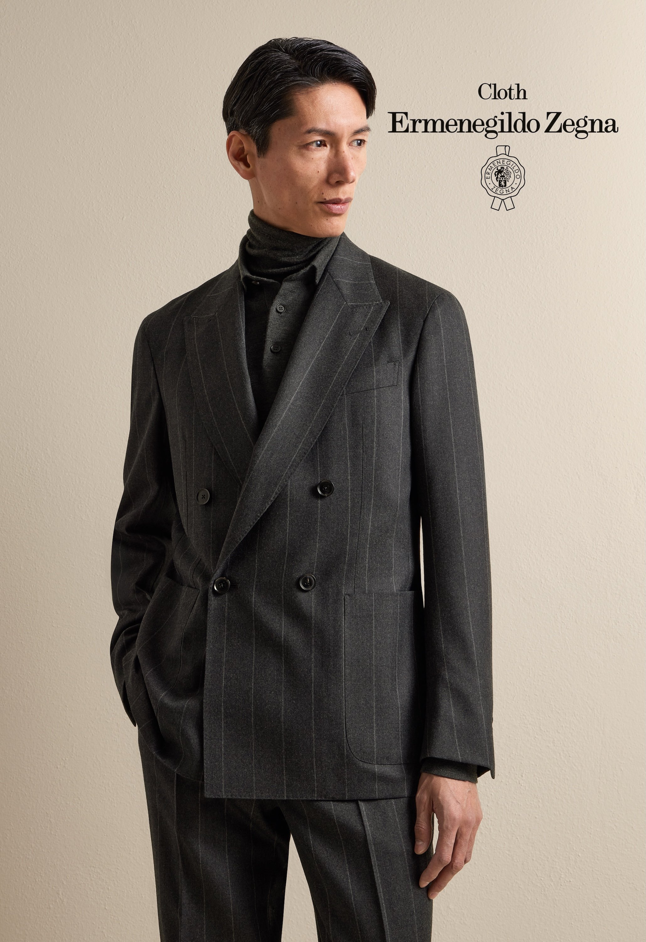 A man wearing a dark pinstripe double-breasted suit with a black turtleneck underneath, standing against a beige wall that has the text 'Cloth Ermenegildo Zegna' and a logo.