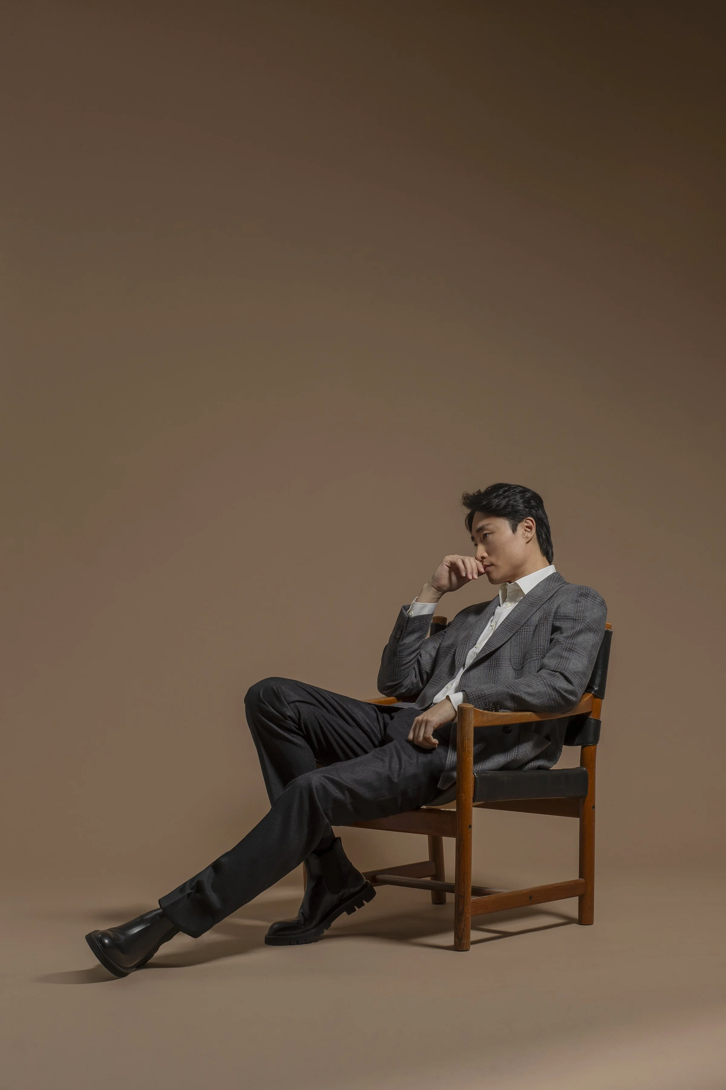 A young man in a Scabal gray plaid blazer, black pants, and black boots sits on a wooden chair against a plain brown background, with his legs crossed and his right hand resting on his chin, appearing contemplative.