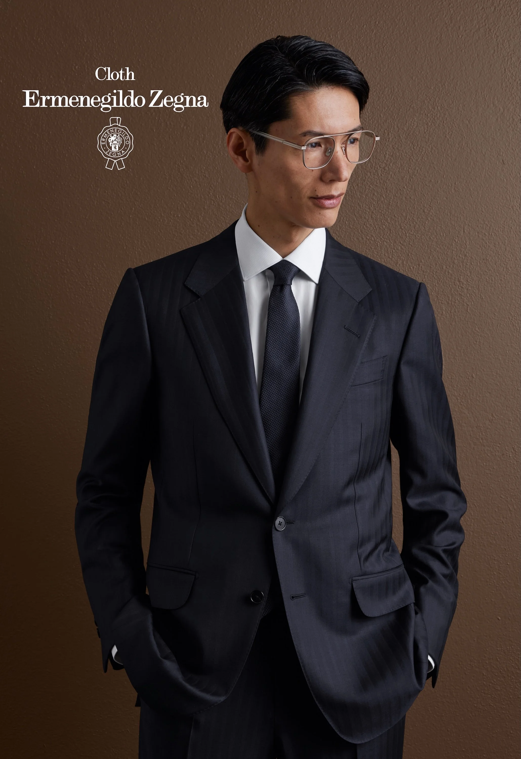 A man wearing a dark pinstripe Zegna suit, white shirt, dark tie, and glasses stands against a brown textured wall, with his left hand in his pocket, looking slightly to the right.