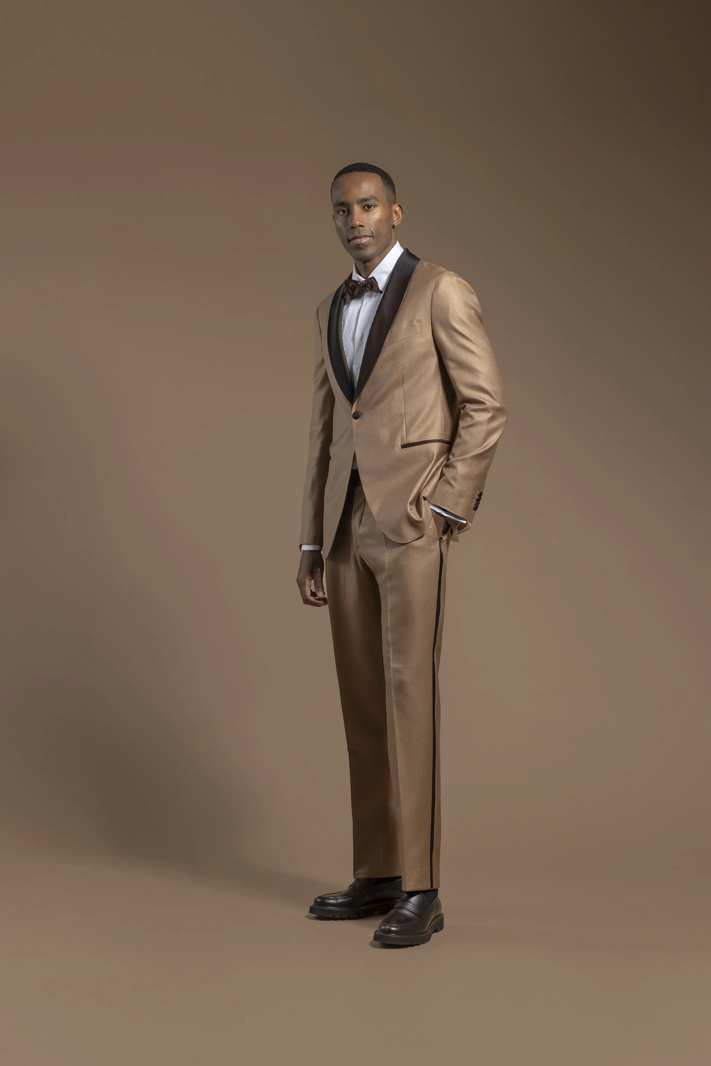 A man wearing a Scabal beige tuxedo with black satin lapels, a white dress shirt, a dark brown bow tie, black dress shoes, and standing against a plain brown background.