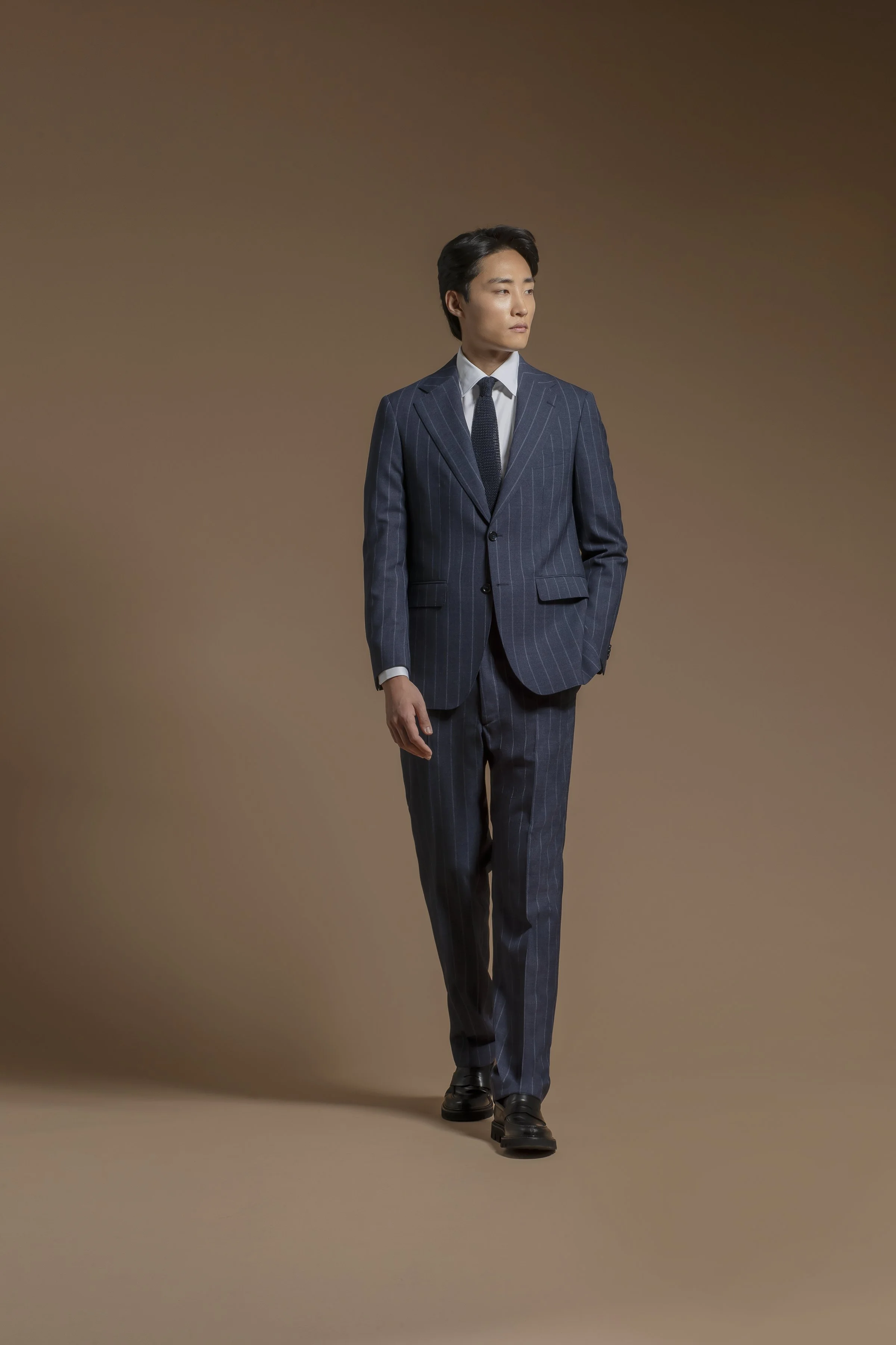 A young man in a dark blue Scabal pinstripe suit, white shirt, and dark tie, walking against a plain brown background.