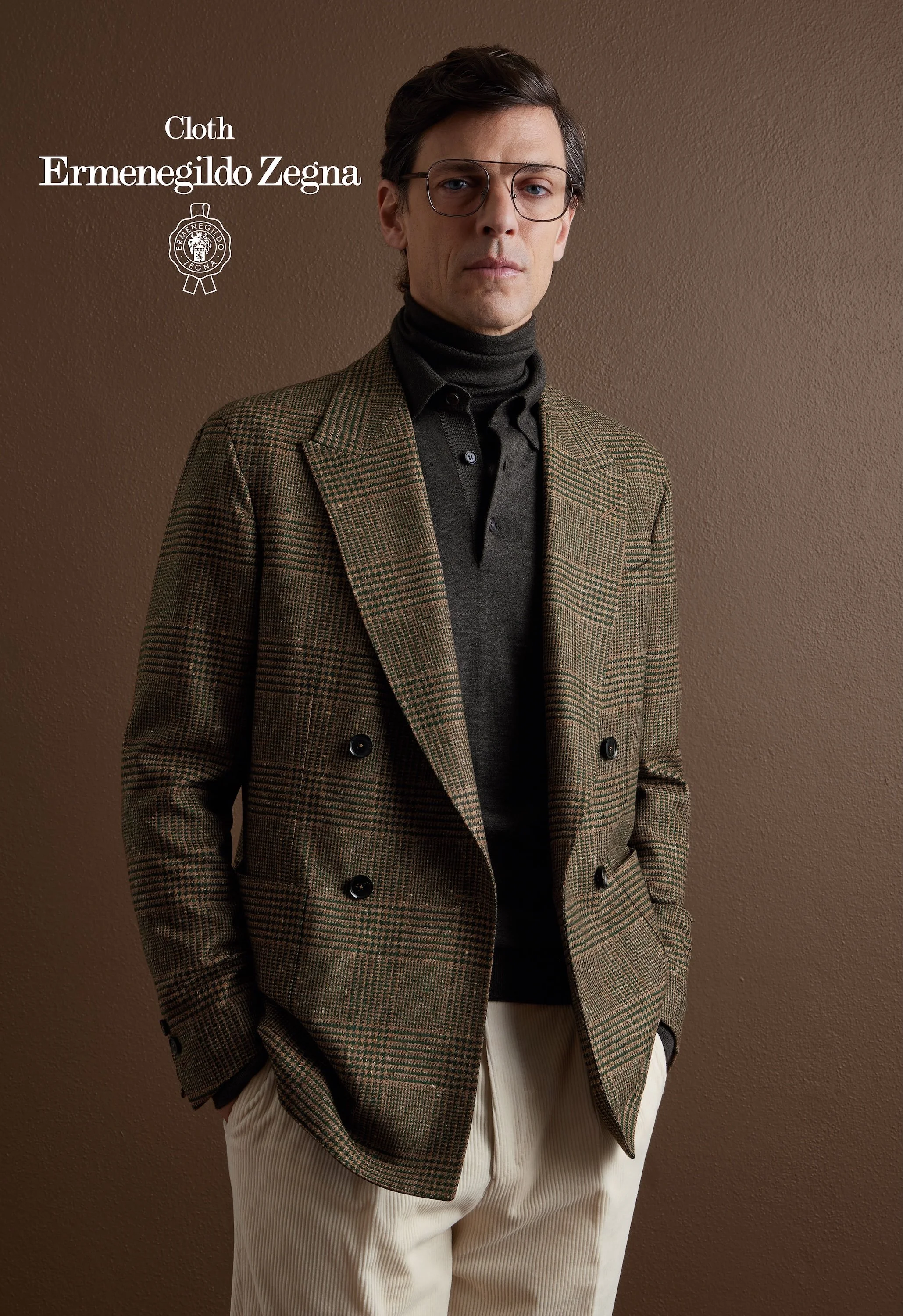 A male model wearing a brown checked Zegna overcoat, black turtleneck, and beige trousers, standing against a brown wall.