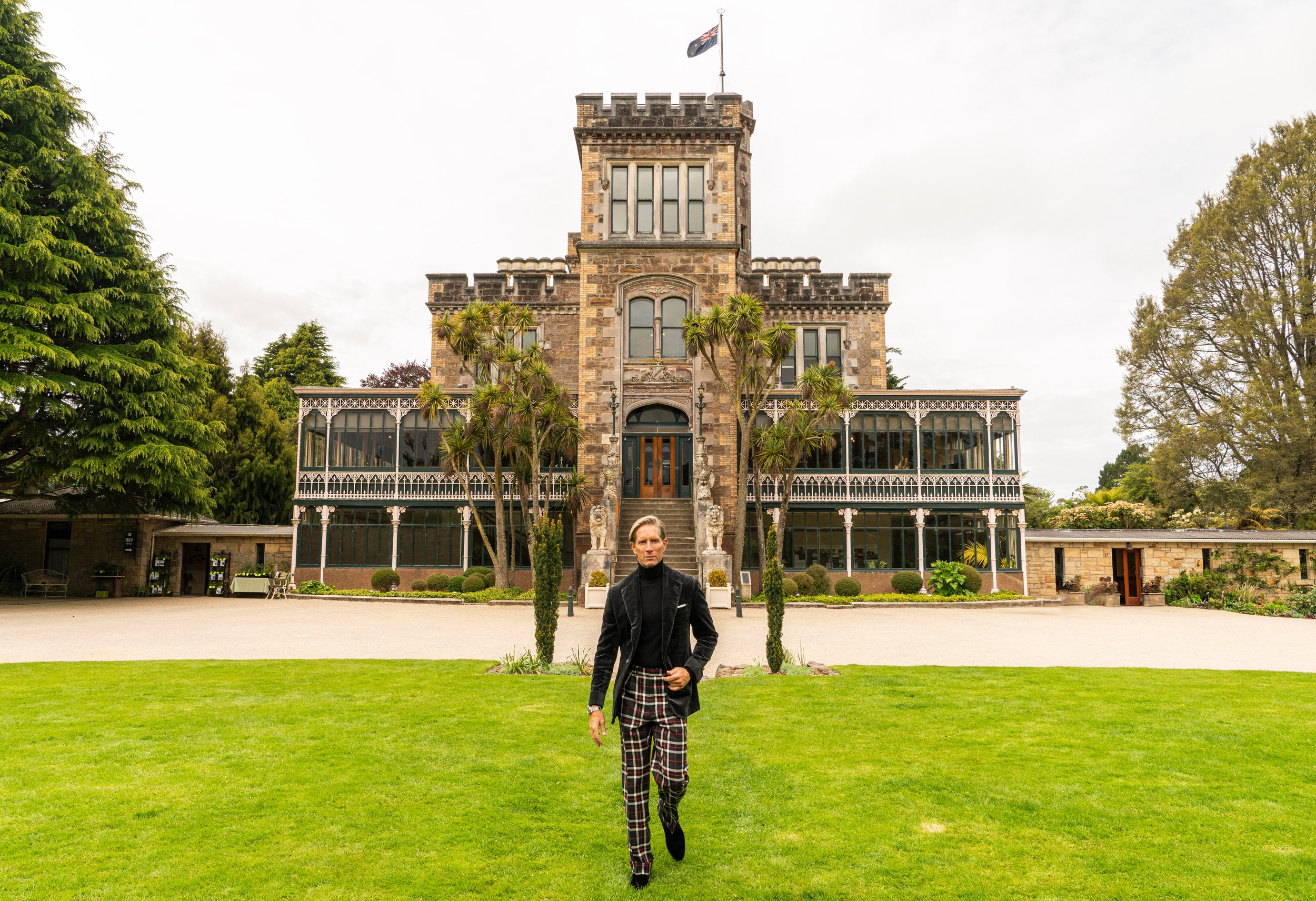 Christian wearing Scarborough Custom Tailoring black jacket and plaid pants walking in front of a historic brick building with ornate detailing and a flag on top, surrounded by green trees and a well-maintained lawn.