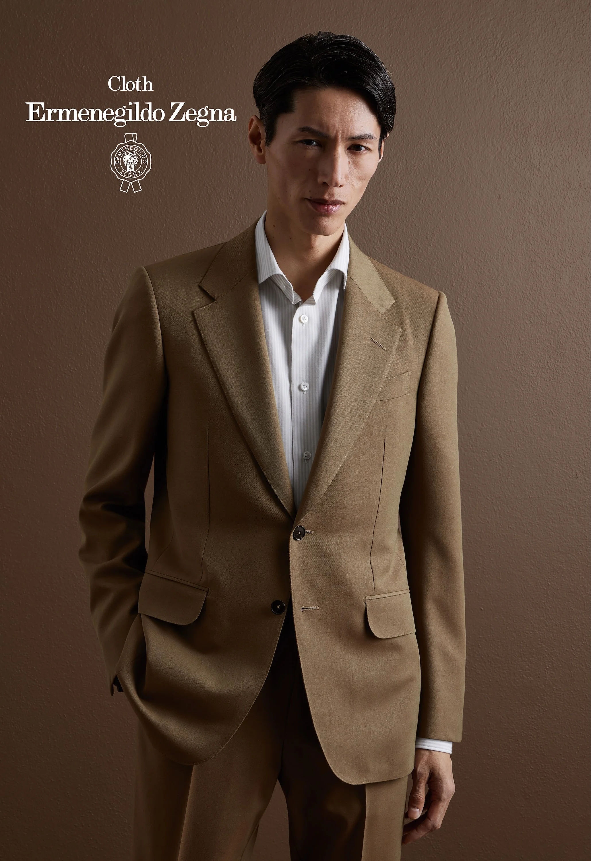 A man wearing a tan Zegna suit with a white shirt standing against a brown wall.