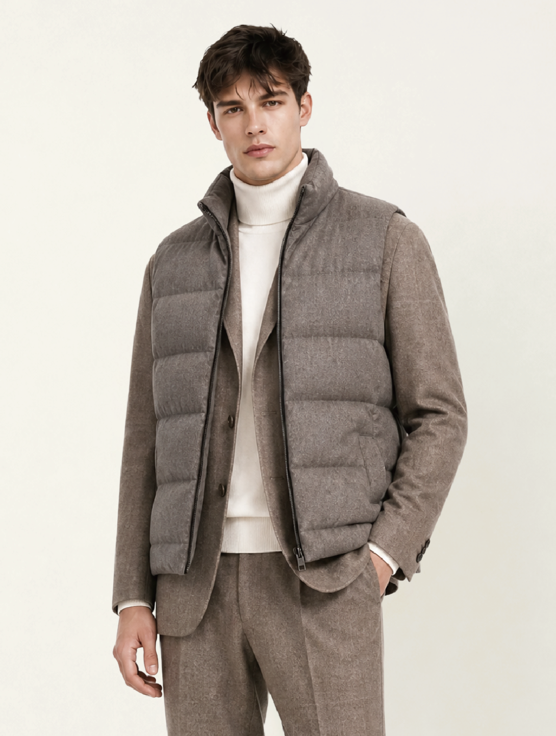 A young man wearing a Scarborough RTW Collection beige suit, a white turtleneck, and a gray quilted vest, standing with his right hand in his pocket against a plain light background.