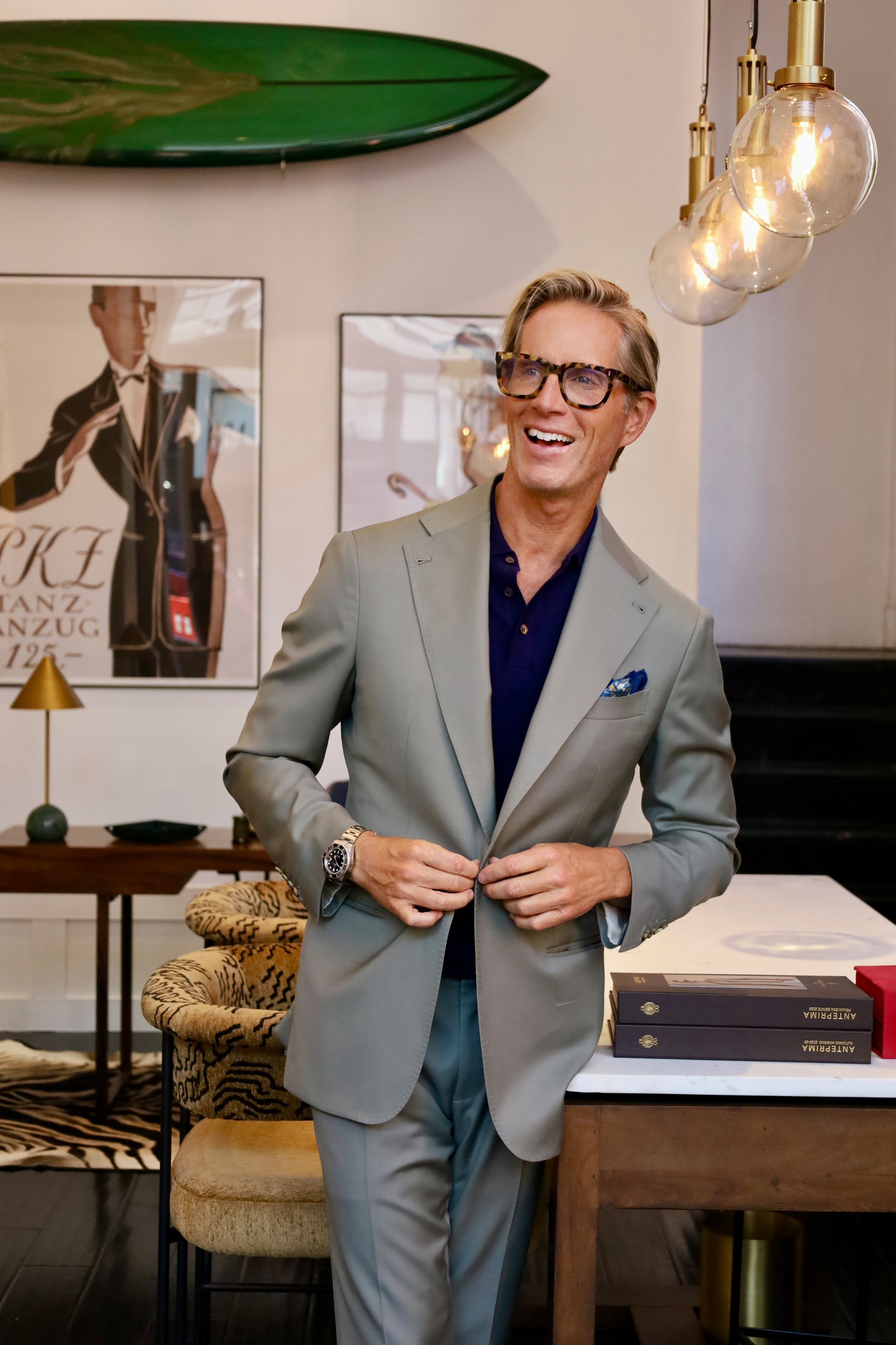 Christian standing indoors at the Scarborough Custom Tailoring showroom in San Francisco, smiling and buttoning his jacket. Modern decor items, including framed artwork, a leopard print chair, a table with books, and hanging light fixtures.