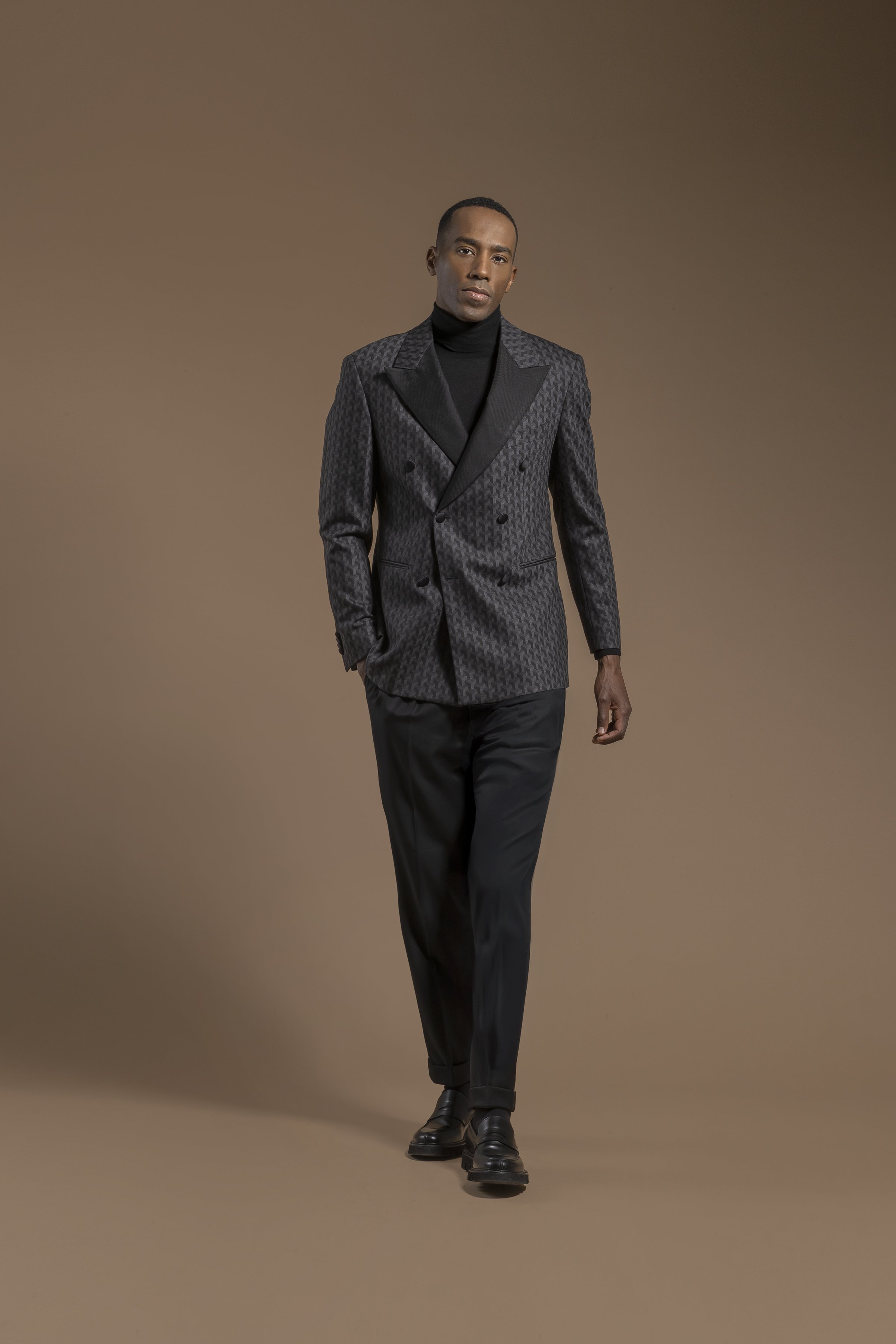 A man walking towards the camera wearing a Scabal dark gray patterned double-breasted blazer over a black turtleneck and black pants, against a plain brown background.