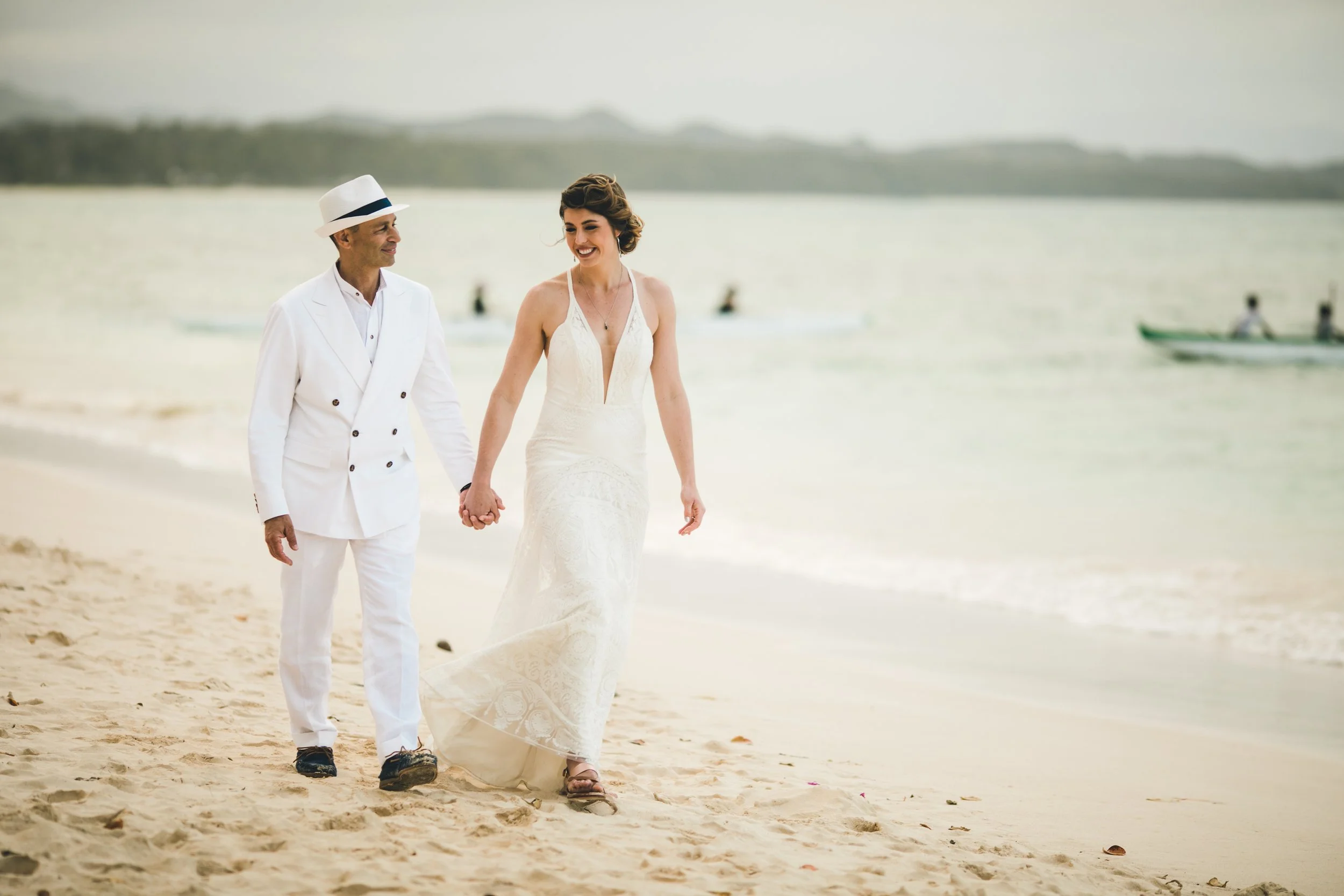 A bride and groom walking hand in hand on the beach, dressed in white Scarborough Custom Tailoring tuxedo wedding attire, smiling and enjoying a moment together with water and boats in the background.