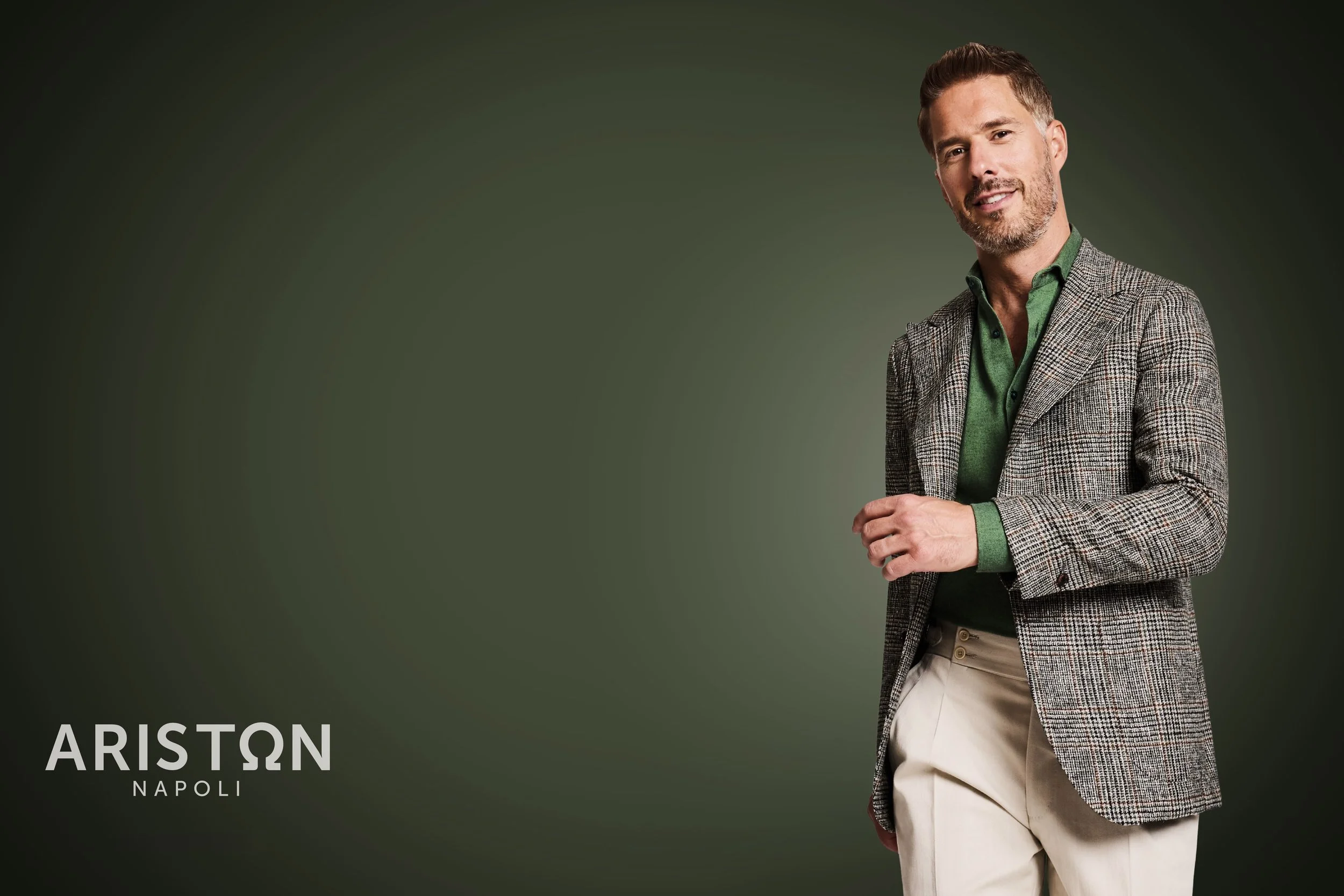 A man with short hair and a beard wearing a green shirt and gray plaid blazer, standing against a dark green background. The bottom left corner features the text 'ARISTON NAPOLI' in white. The man is smiling with his arms crossed.