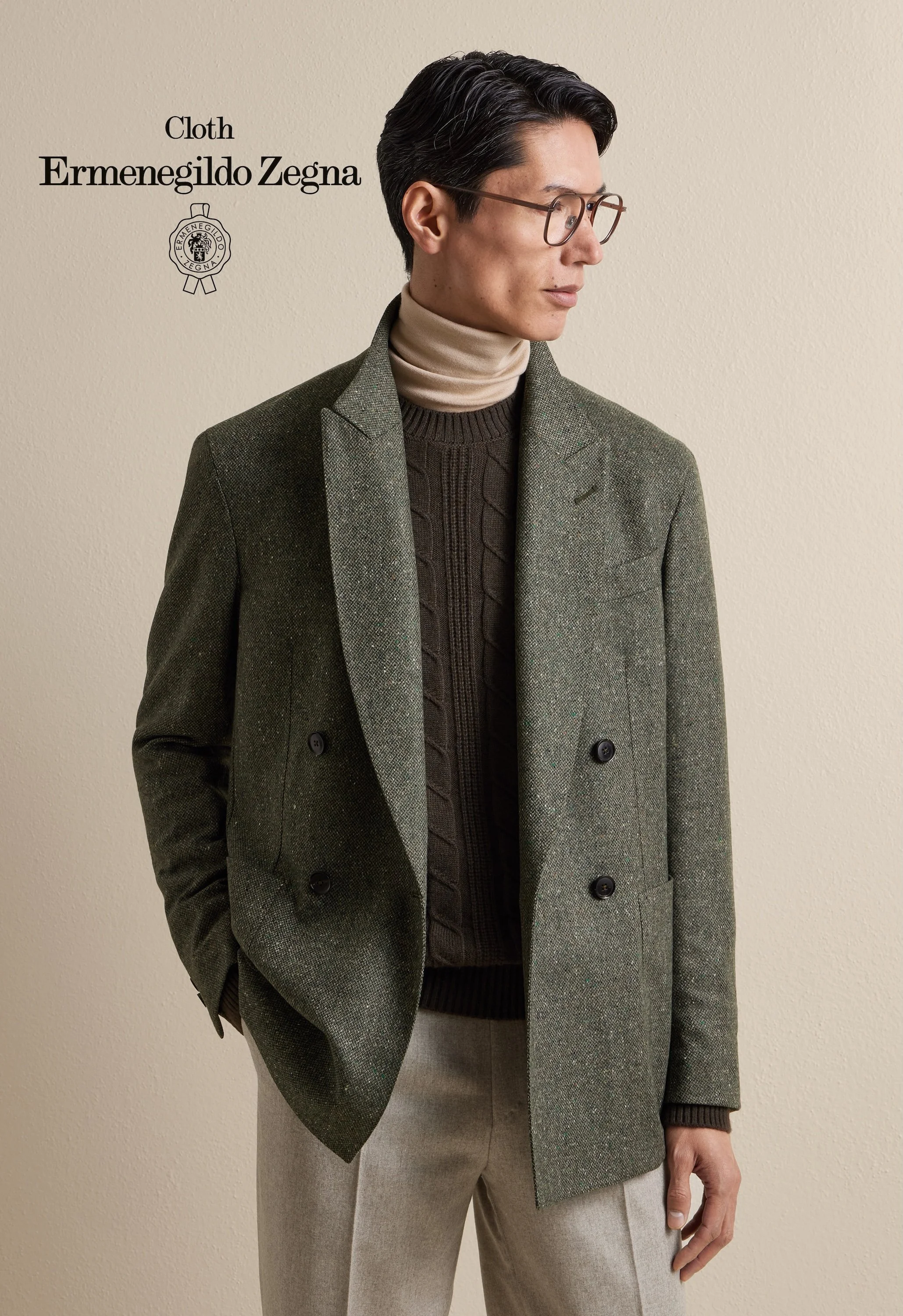 A man wearing glasses, a beige turtleneck, a dark brown sweater, a green checked Zegna overcoat, and beige pants, standing against a plain beige wall.