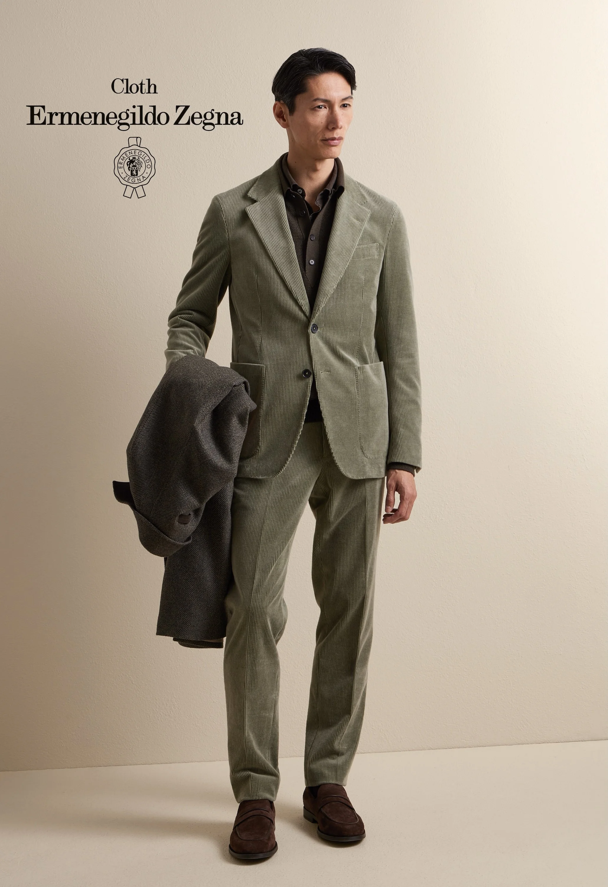 A man in a beige corduroy suit holding a dark gray blazer, standing against a plain beige wall with text indicating the clothing is from Ermenegildo Zegna.