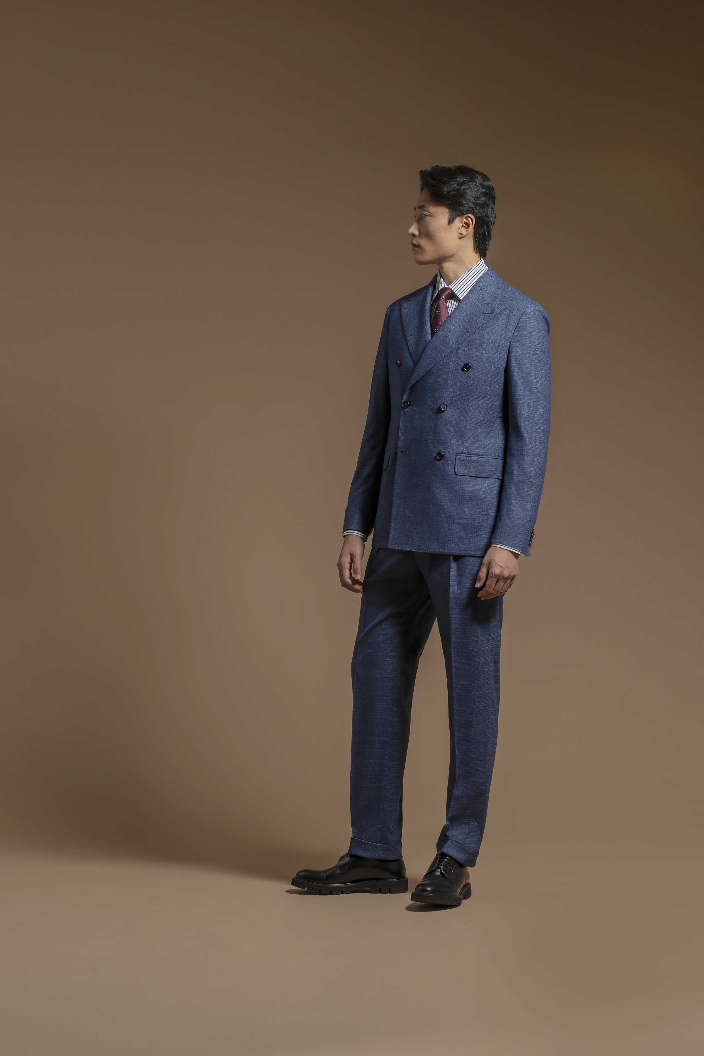 A man dressed in a Scabal double-breasted blue suit, striped shirt, and burgundy tie, standing against a plain brown background. He is looking to his right with a serious expression.