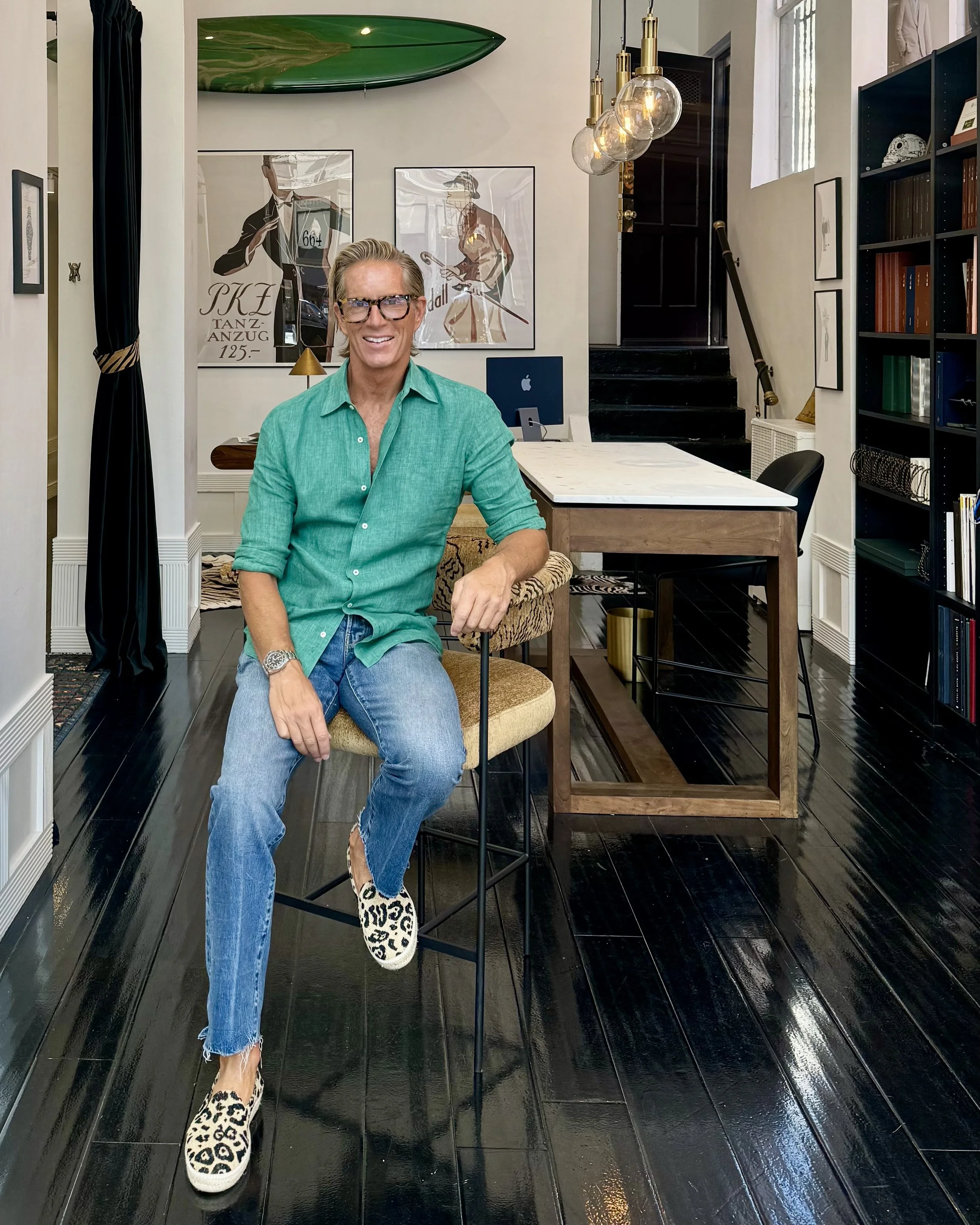 Christian Gardner with blonde hair, eyeglasses, and a green shirt sitting in the Scarborough Custom Tailoring showroom with dark wood floors, black stairs, and artsy decor, including framed artwork, a surfboard.