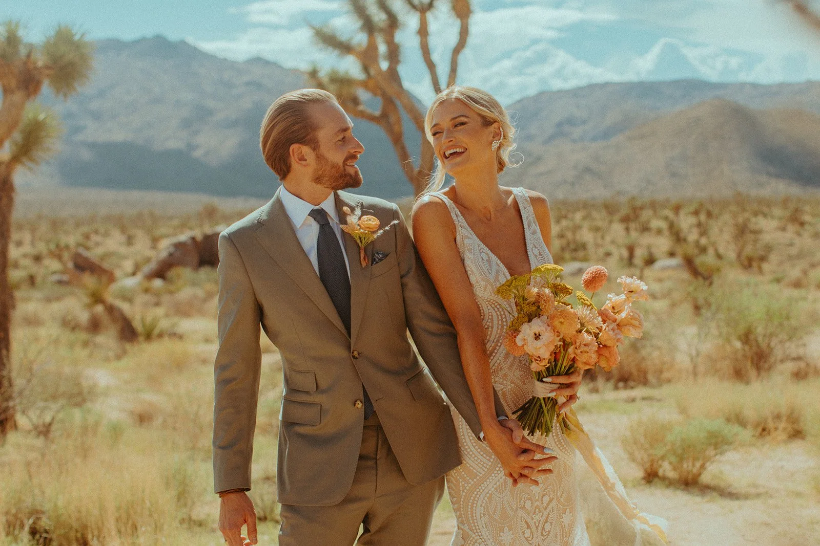 A smiling bride and groom wearing Scarborough Custom Tailoring suit holding hands and a bouquet of flowers in a desert landscape with mountains, Joshua trees, and blue sky.