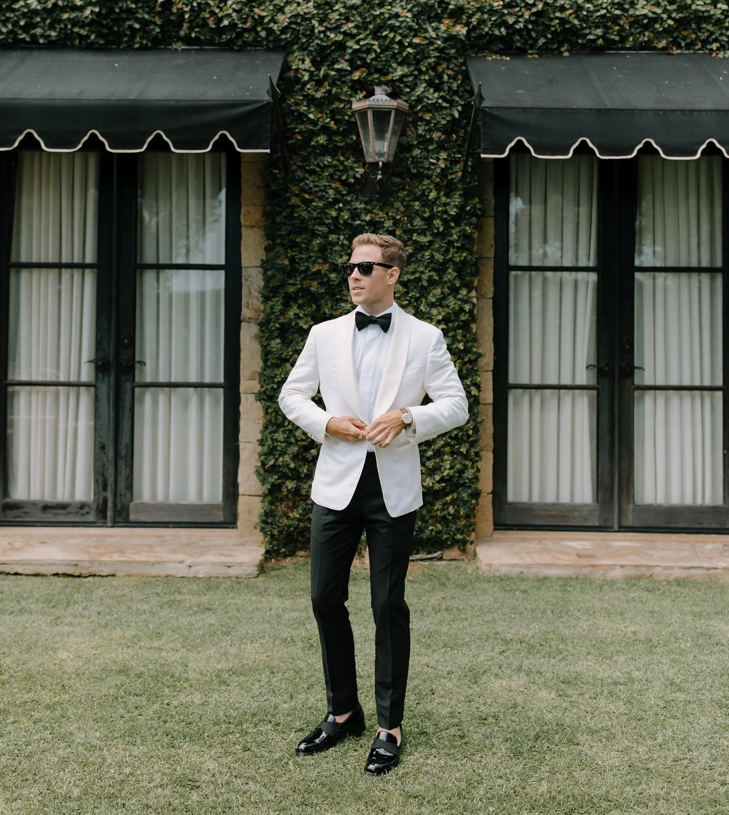 A groom wearing a Scarborough white tuxedo jacket, black pants, black buckle shoes, a bow tie, and sunglasses standing in front of a building with black doors and windows covered by white curtains, surrounded by green grass and a leafy background.