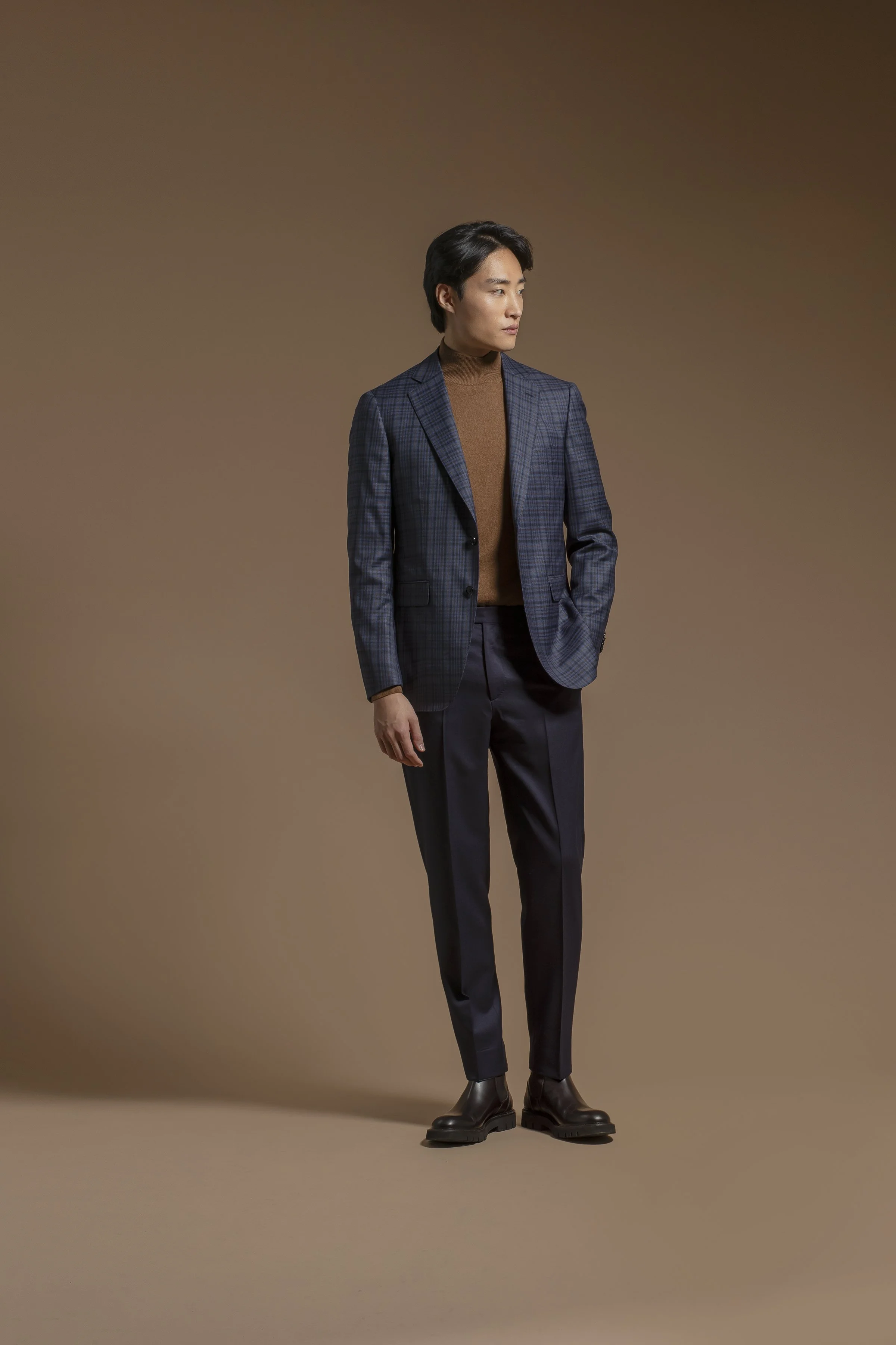 A man dressed in a Scabal dark plaid blazer, tan turtleneck, dark pants, and black boots, standing against a brown gradient background, looking to the side with hands in pockets.