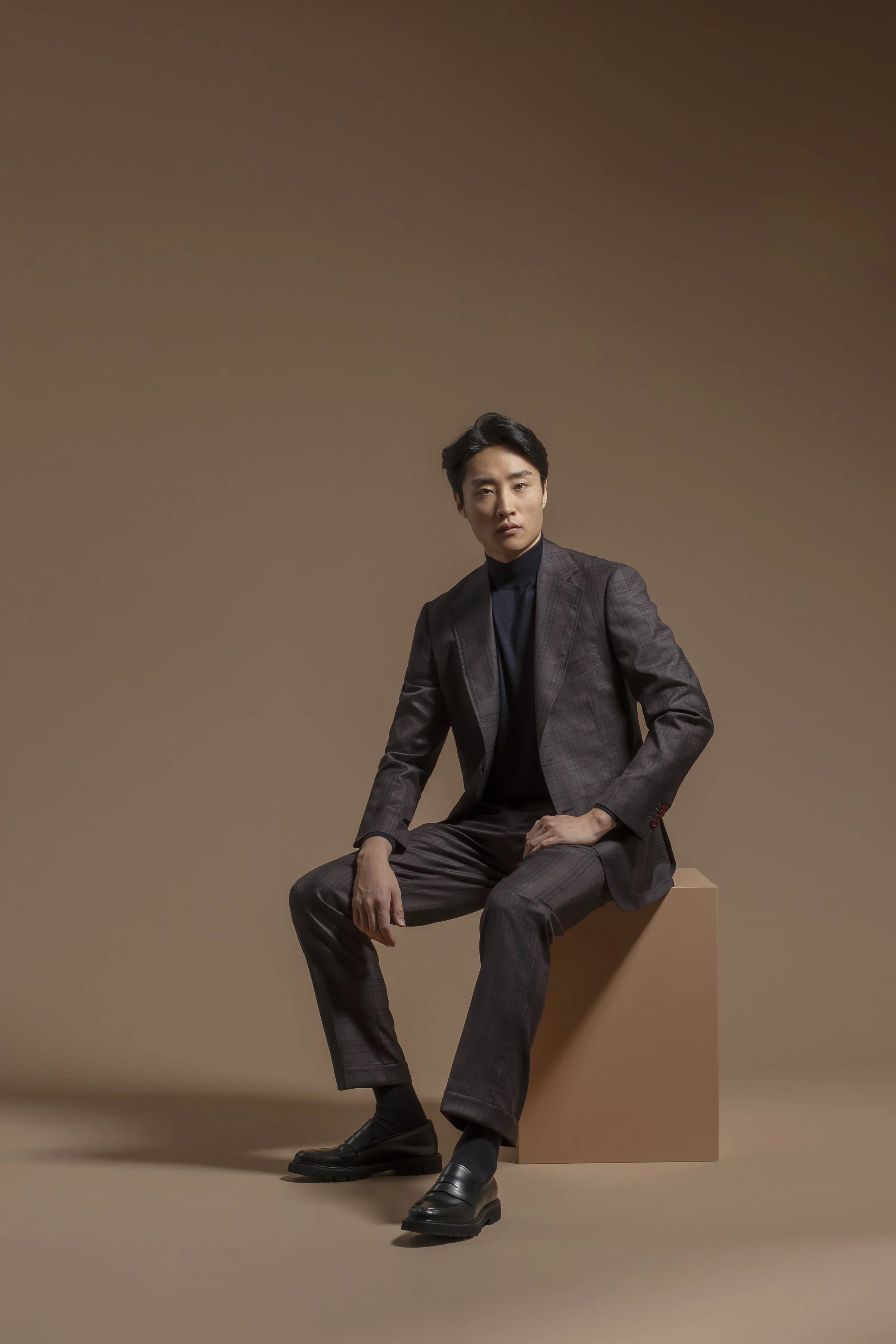 A man with black hair sitting on a small table against a plain beige background. He is wearing a Scabal dark plaid suit with a black turtleneck top and black shoes.