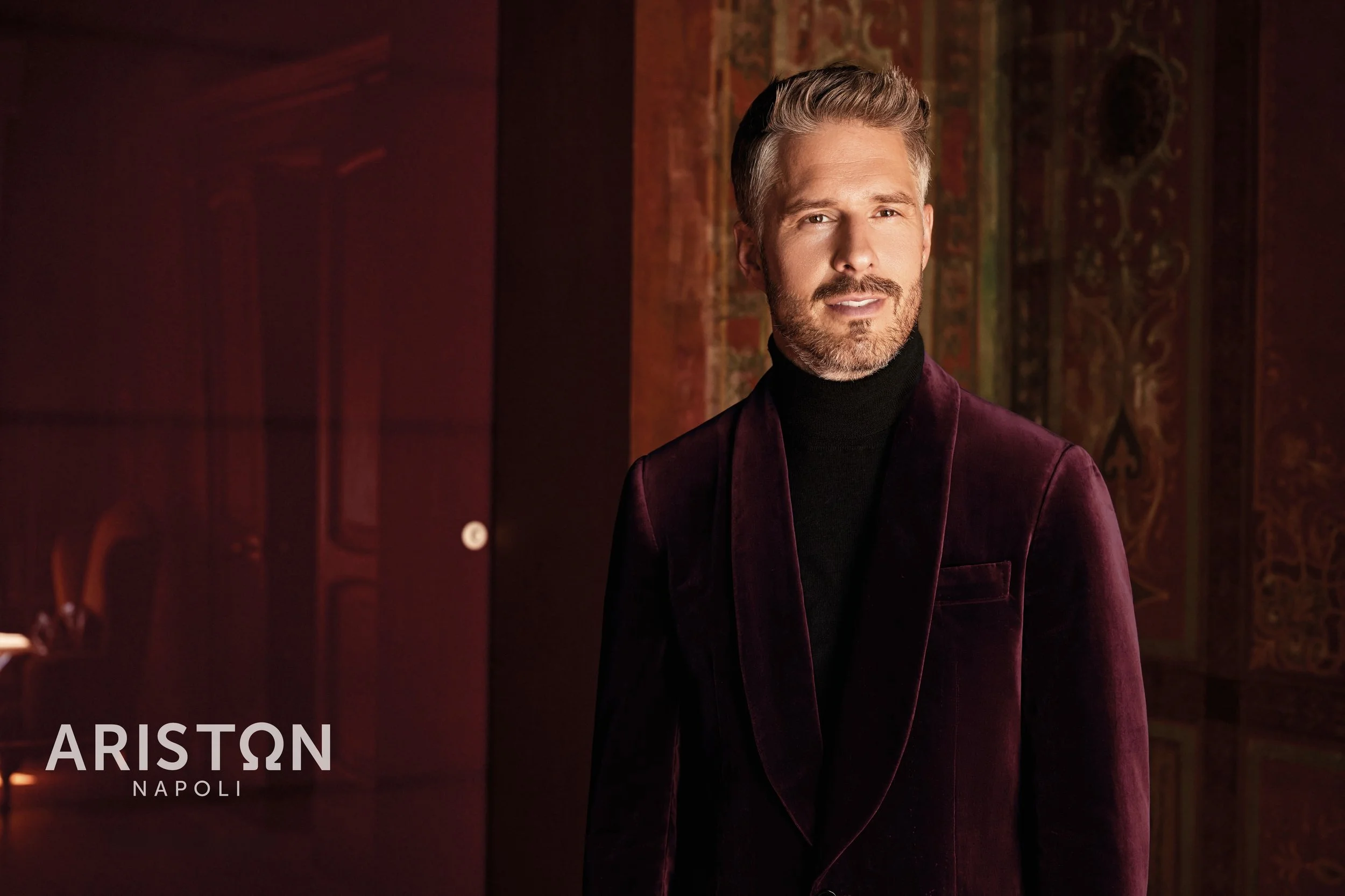 A man with blonde hair and a beard, dressed in an Ariston dark turtleneck and a maroon blazer, standing in front of a patterned background with warm lighting.