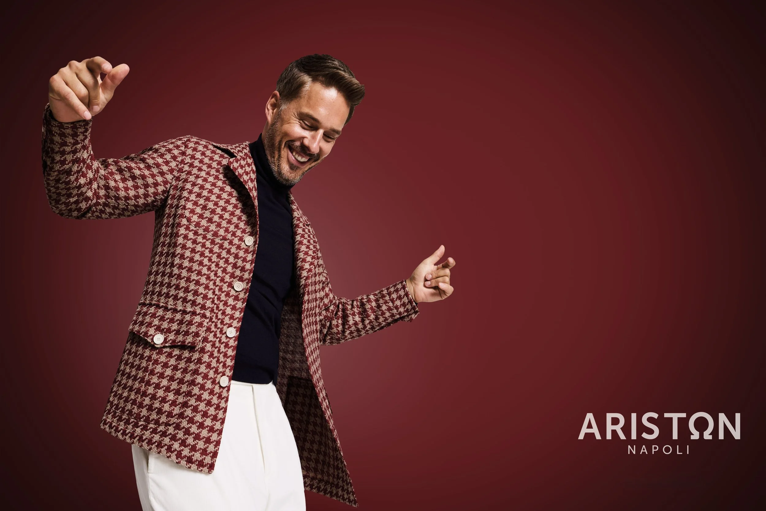 A man with light skin, facial hair, and short brown hair, wearing a red and white houndstooth patterned blazer over a black shirt and white pants, smiling and dancing against a dark red background. The text 'ARISTON NAPOLI' is visible in the bottom r