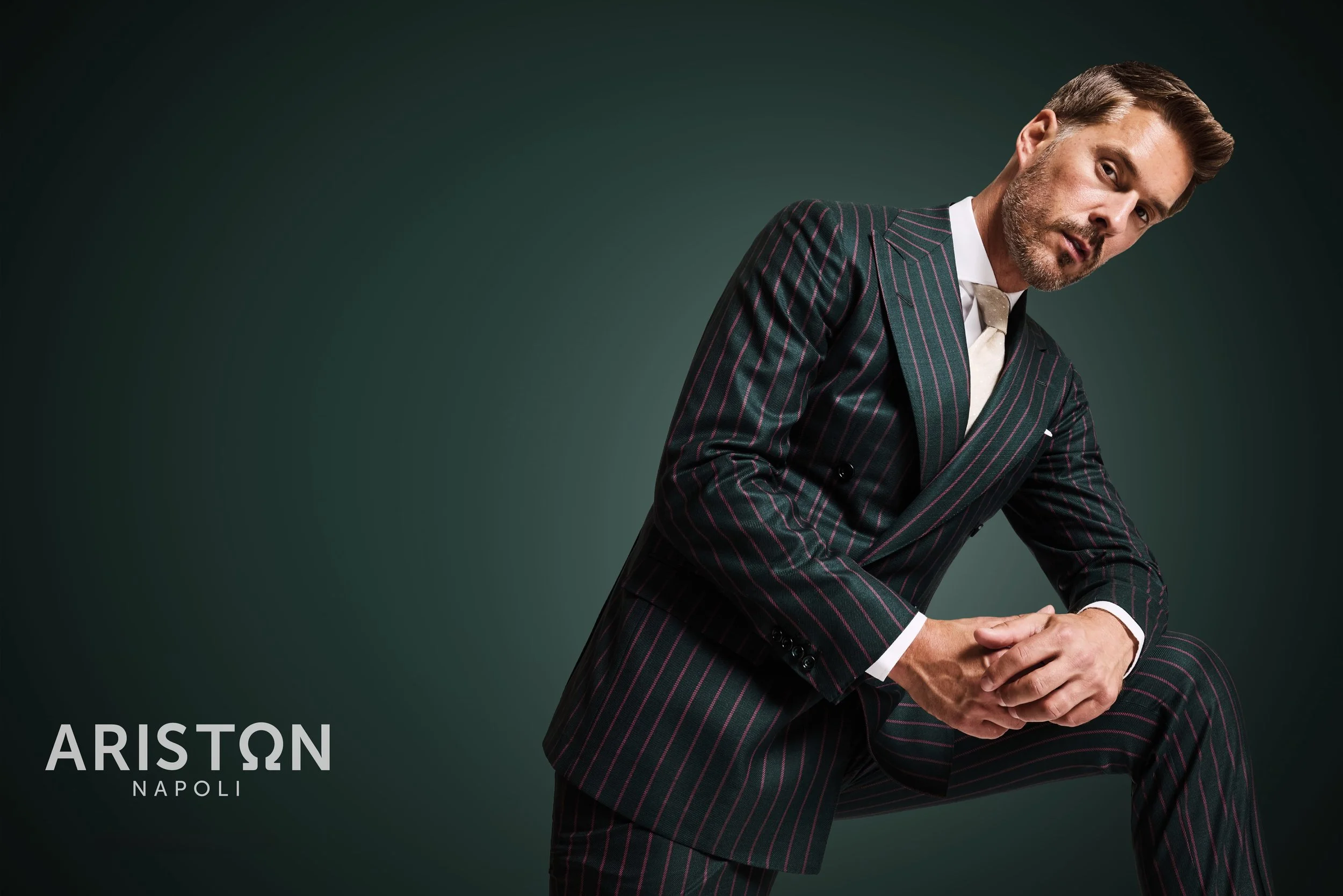 Man in a dark pinstripe suit with a white shirt and beige tie, posing against a dark green background, looking at the camera with a serious expression. The text 'ARISTON NAPOLI' appears at the bottom left corner of the image.