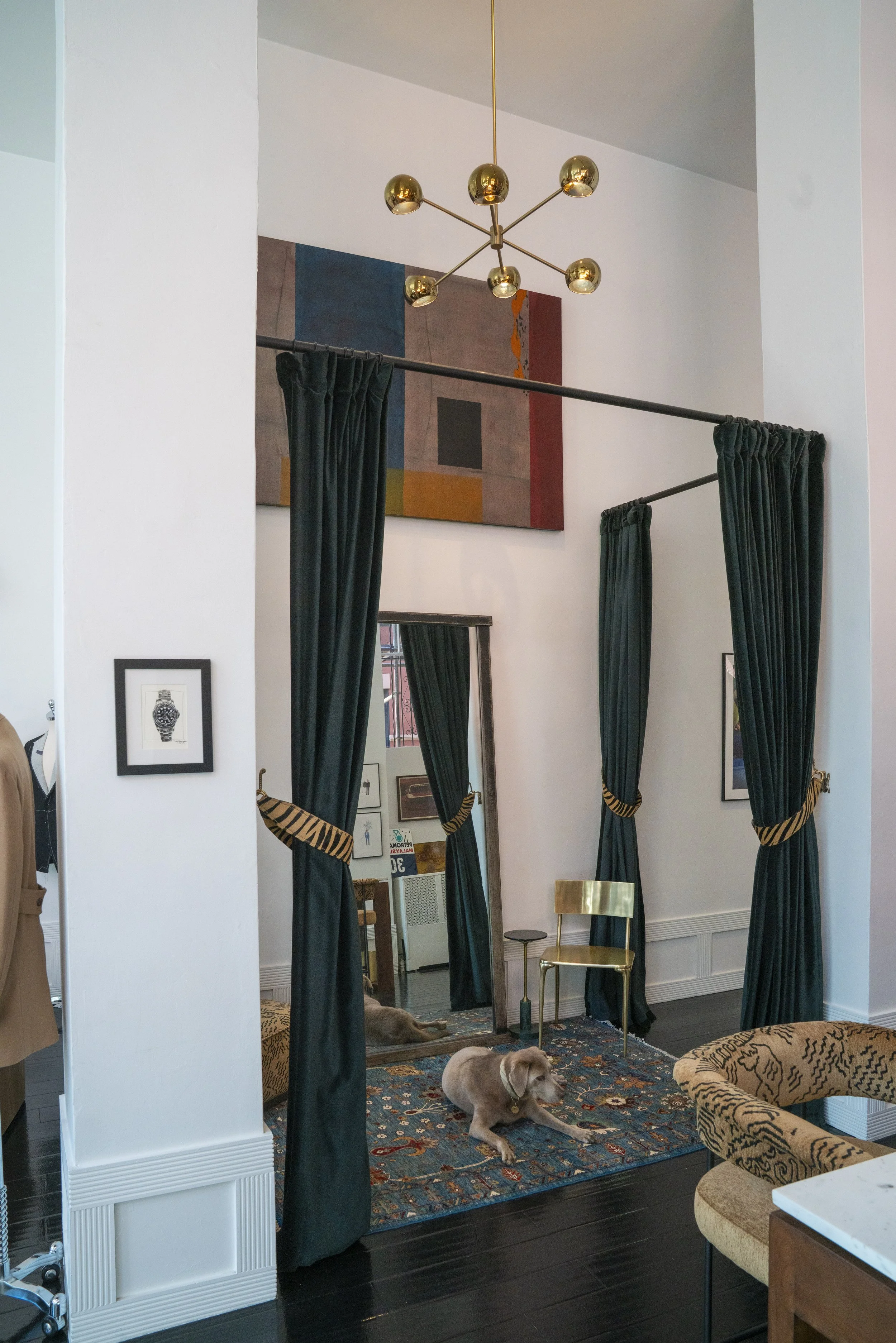 Dressing room of Scarborough Custom Tailoring showroom in San Francisco with dark wood floors, a colorful patterned rug, gold chairs, black curtains, a modern chandelier with gold sphere bulbs, framed pictures on the wall and a floor mirror.