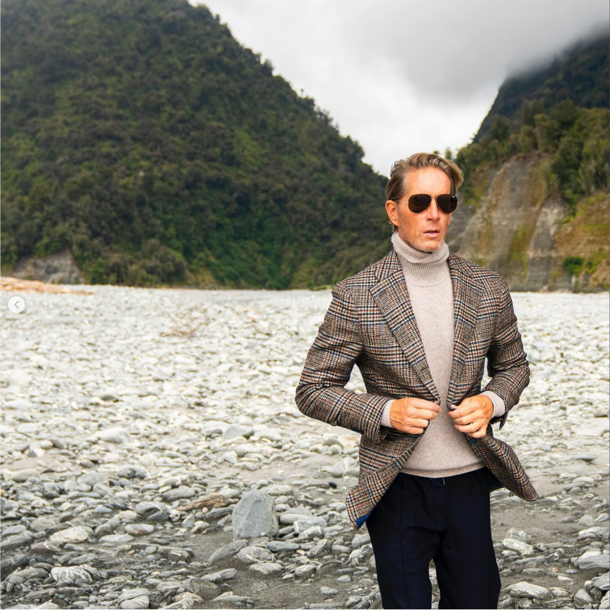 Christian wearing sunglasses, a plaid blazer, and a turtleneck, standing on a rocky riverbank with mountains and cloudy sky in the background in New Zealand.