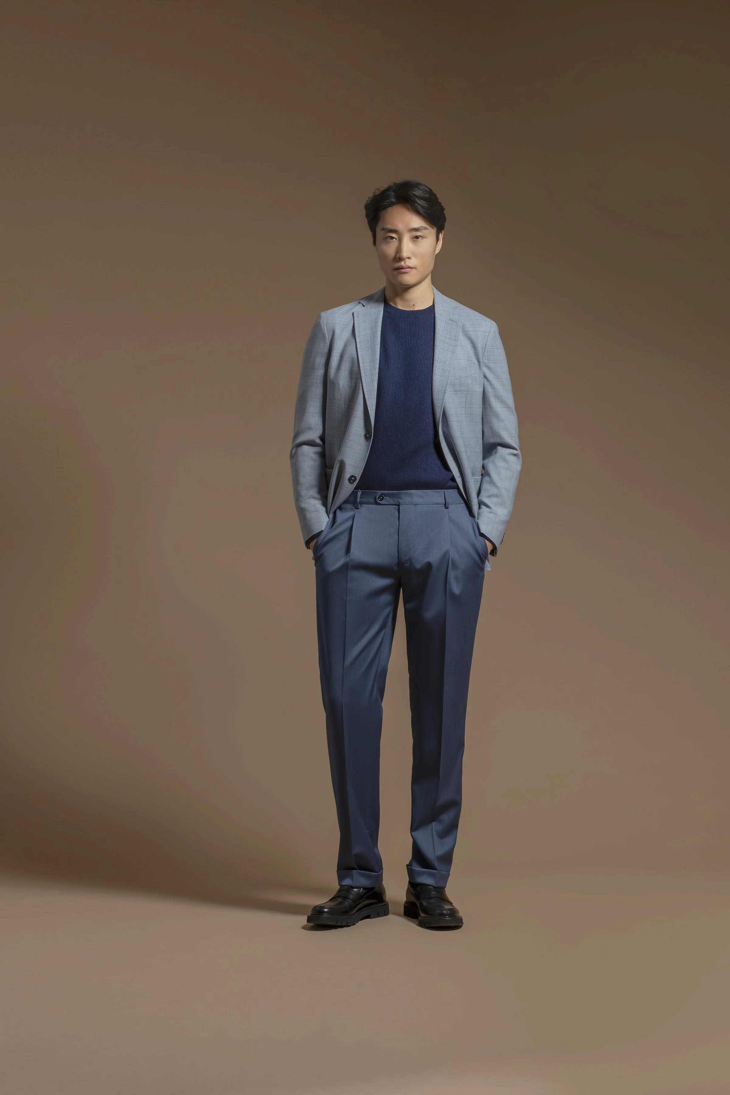 A man in a Scabal blue suit, blue shirt, and black shoes stands against a plain brown background with hands in his pockets.