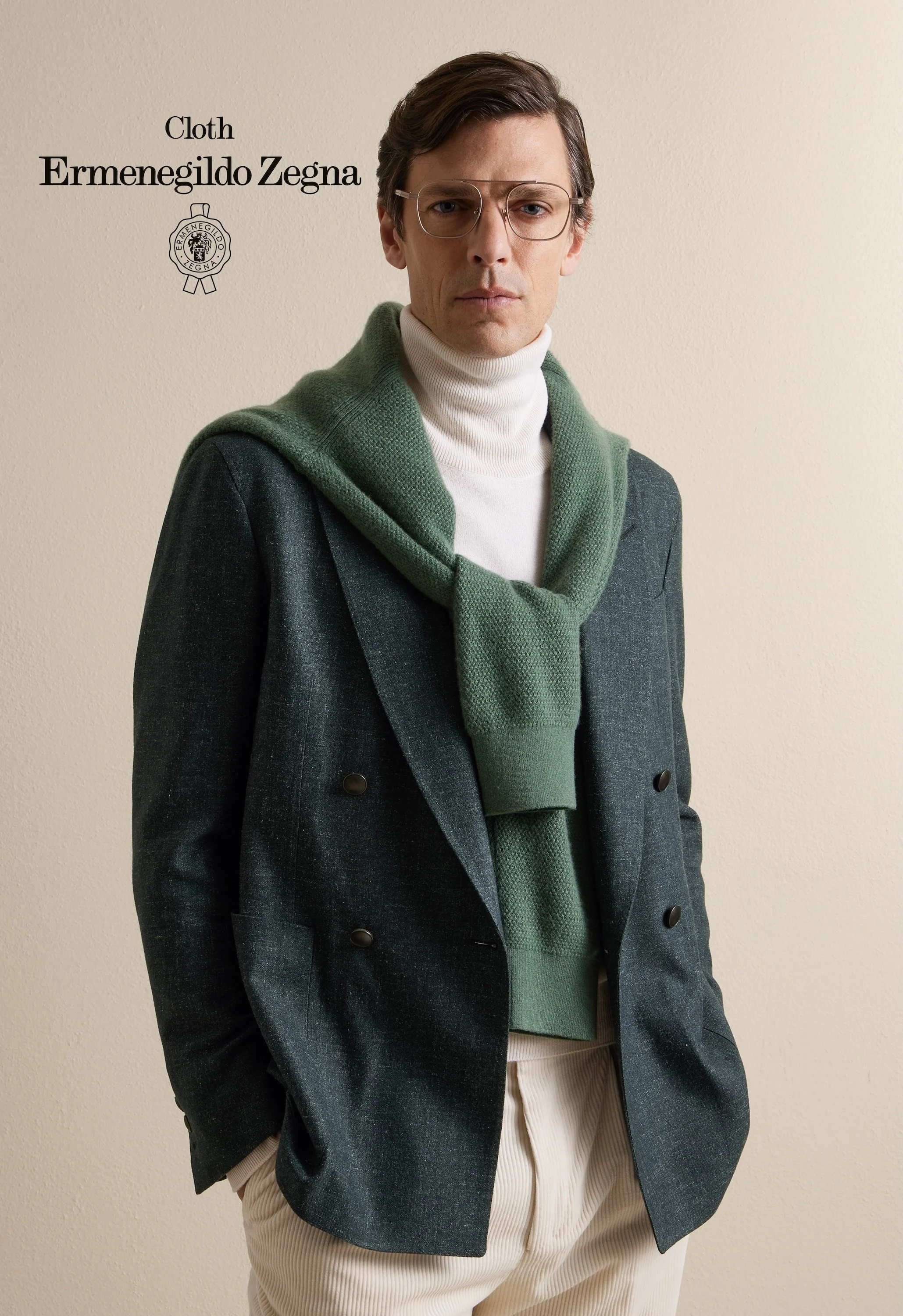 A man wearing glasses, a white turtleneck, a green scarf, a dark gray coat, and beige pants stands against a plain beige wall. Text on the wall reads "Cloth Ermenegildo Zegna" with a logo of a bee.