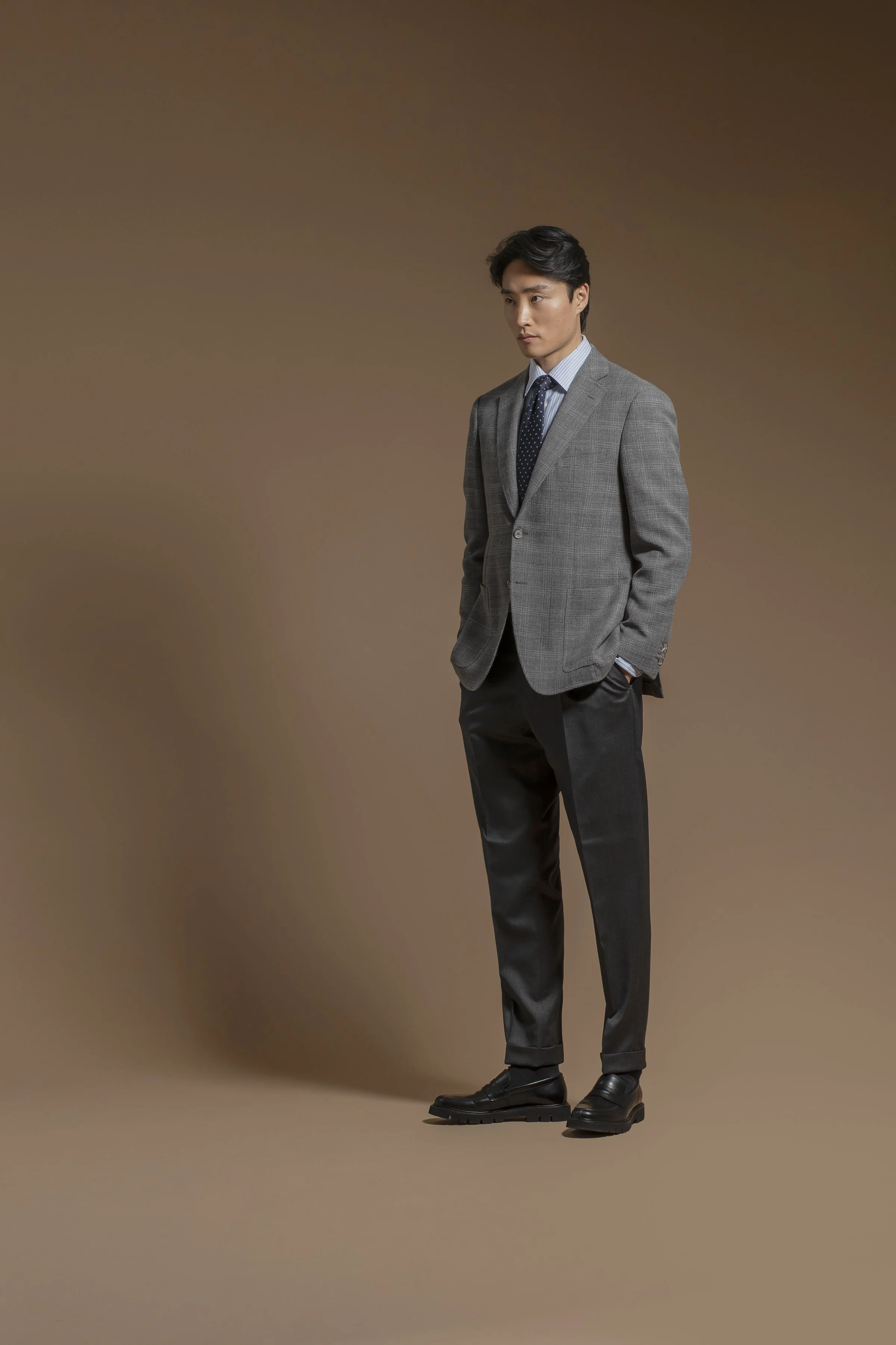 A young man in a Scabal gray suit with a blue shirt and a dark chevron patterned tie standing against a plain tan background. He has dark hair and is looking off to the side with his hands in his pockets.