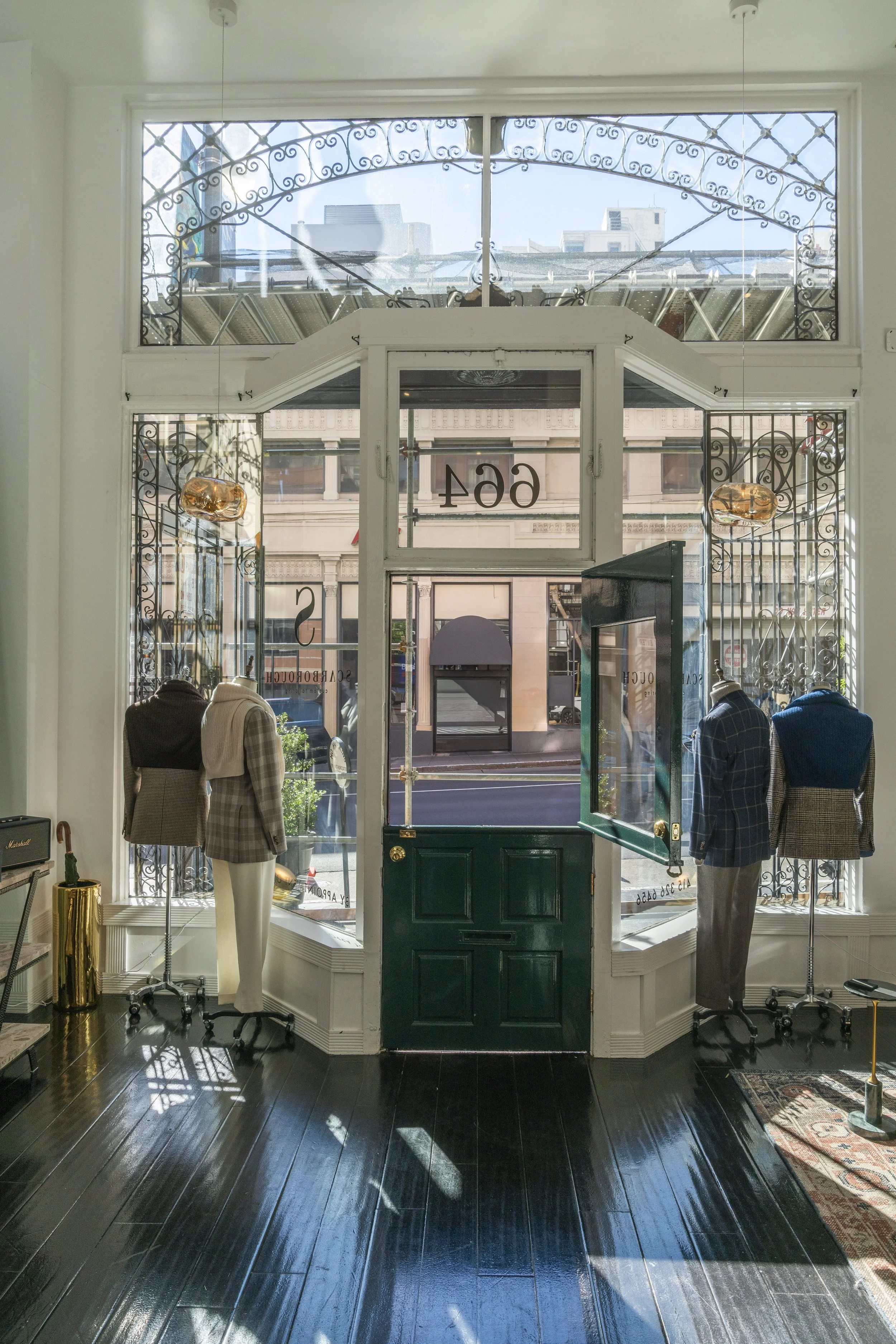 View from the showroom looking out the front window with three mannequins dressed in men's suits, a green door, and a city street outside.