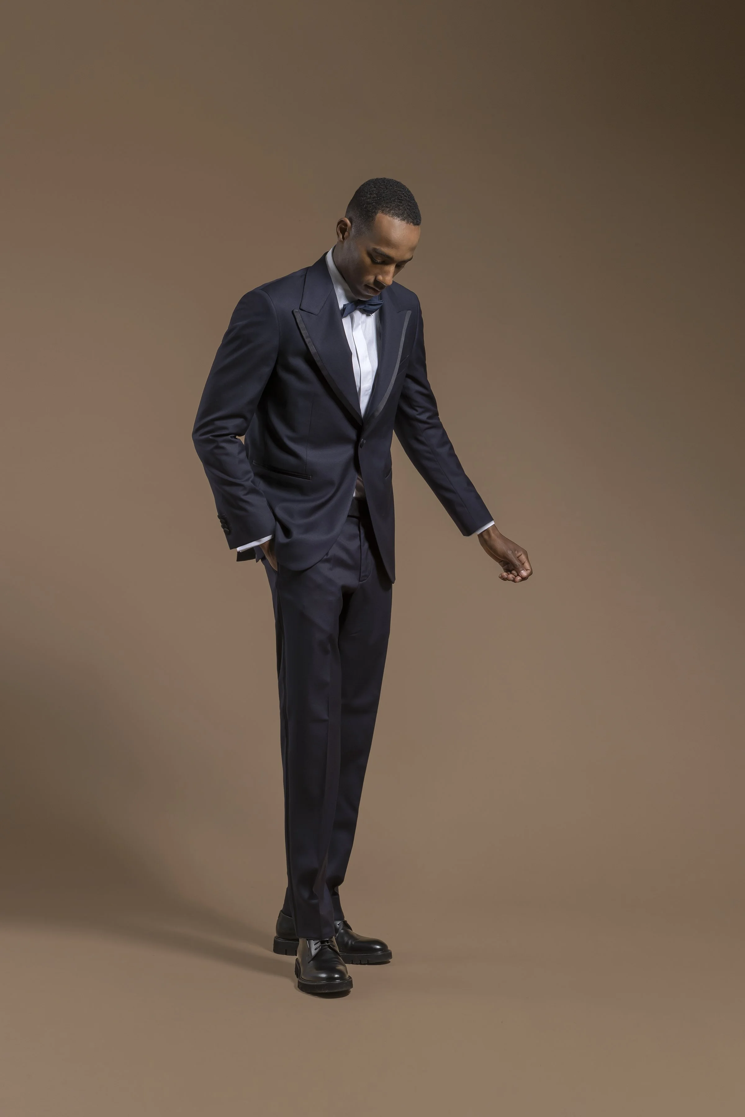 A man in a dark navy Scabal suit with a white shirt and blue bow tie, standing against a brown gradient background, looking down with a relaxed posture.