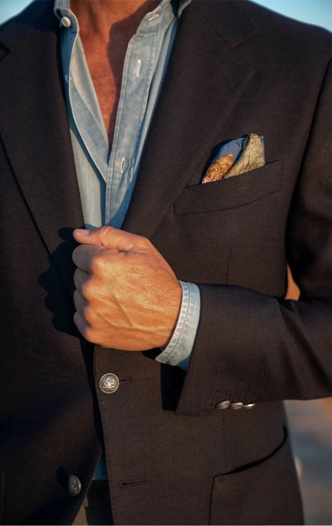 A light blue button-down chambray shirt worn open at the collar under a dark textured sport coat with a paisley pocket square.