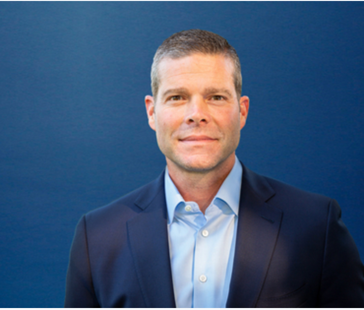 Portrait of a man in a navy blue suit and light blue shirt against a solid blue background.