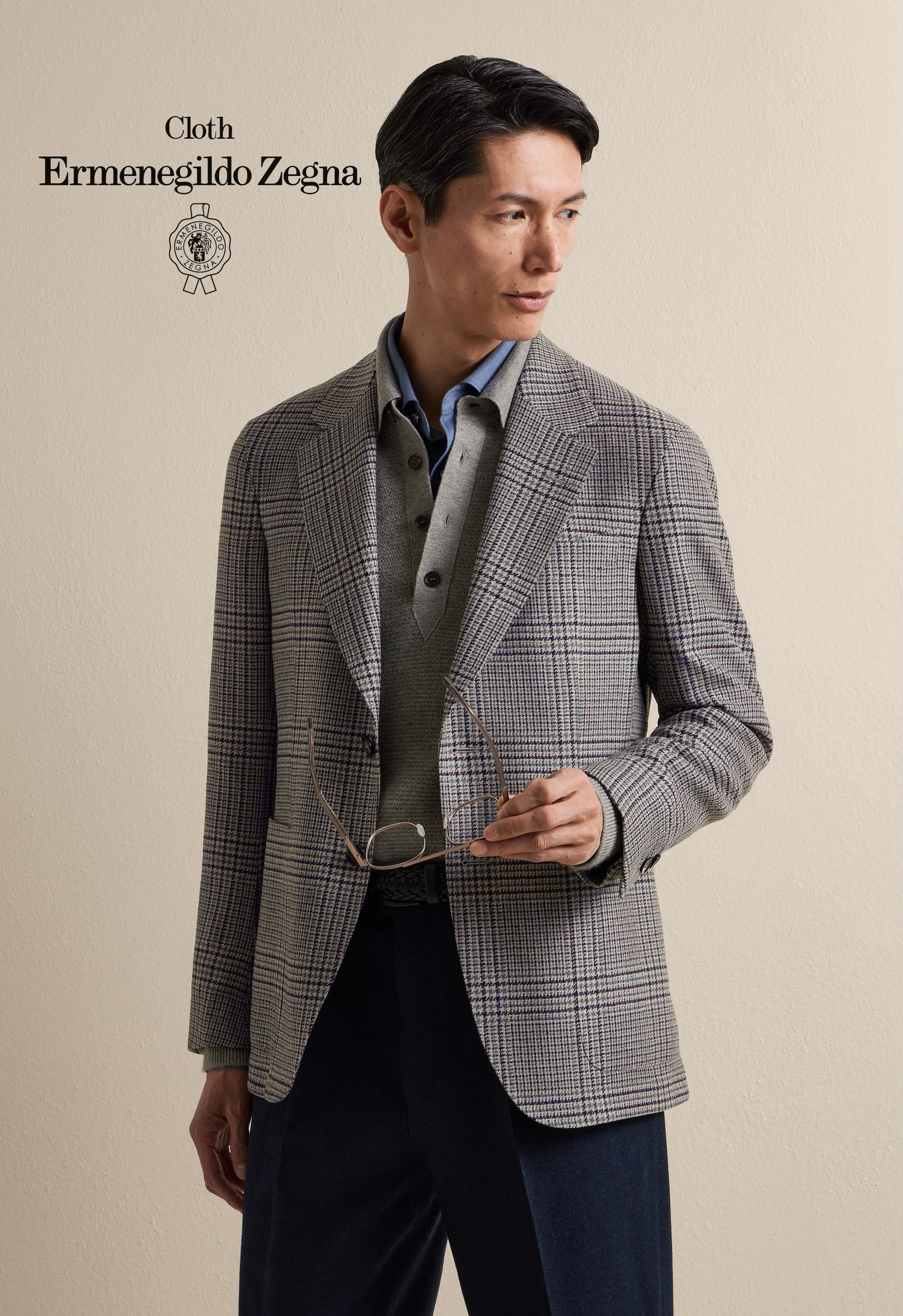 A man wearing a checked Zegna blazer, grey vest, and blue shirt holding glasses, standing against a beige wall.