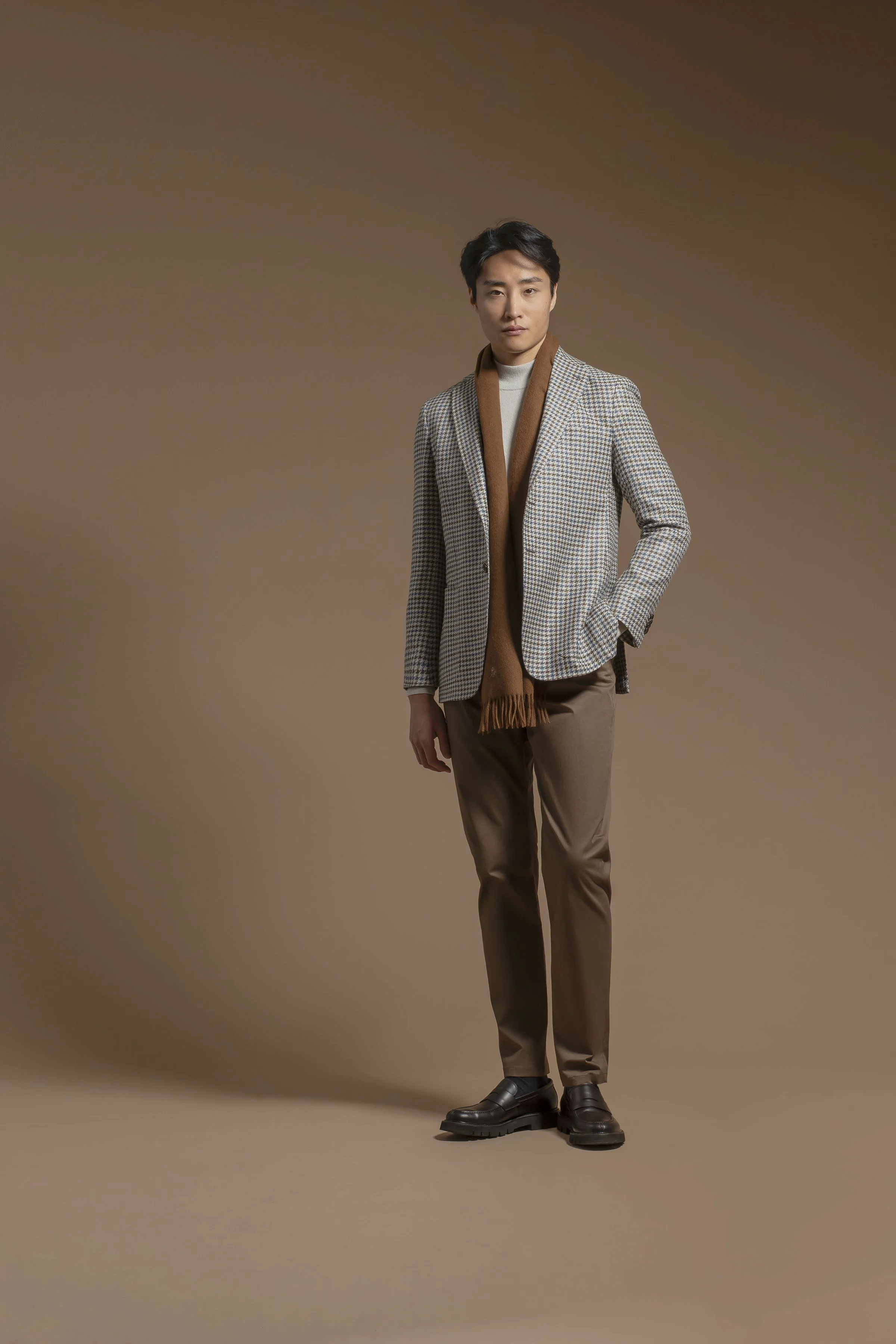 A young man with black hair dressed in a Scabal grey checked blazer, beige pants, and black shoes, standing against a brown background. He has a neutral expression and is posing with one hand in his pocket.