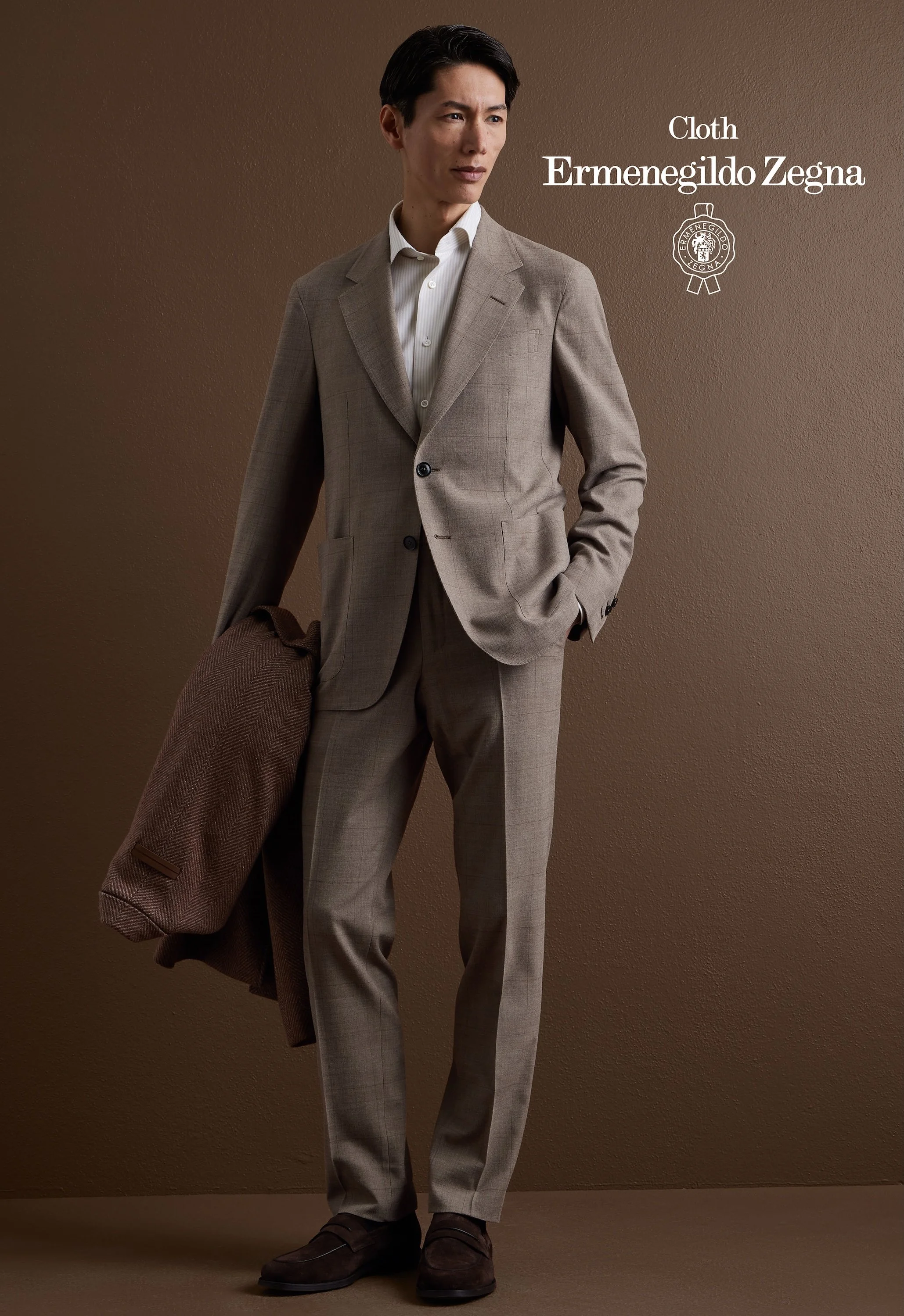 A man in a light brown Zegna suit with a white shirt holding a brown coat, standing against a plain brown background.