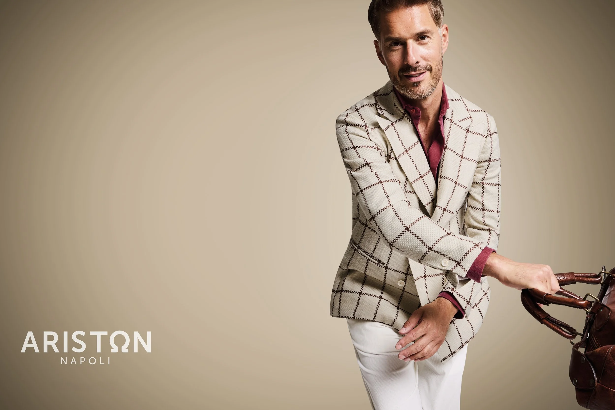 Man in a cream-colored suit with a windowpane pattern, pink shirt, and white pants, holding a vintage camera. The background is plain beige. Logo of Ariston Napoli in the lower-left corner.